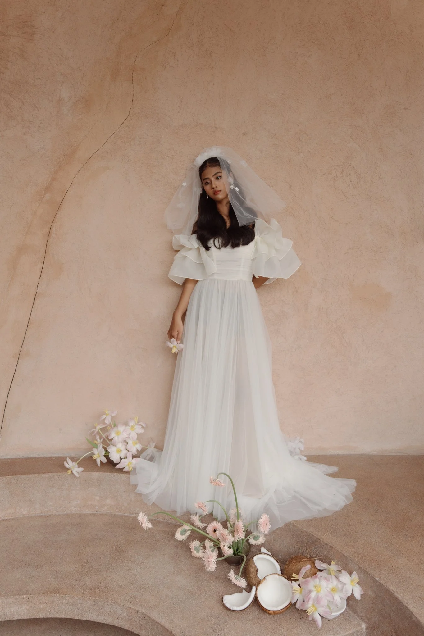 A woman in a white dress with puffed sleeves and a veil stands against a textured beige wall, holding flowers. There are pink and white flowers on the ground, along with broken coconut shells and a small white candle near her feet.