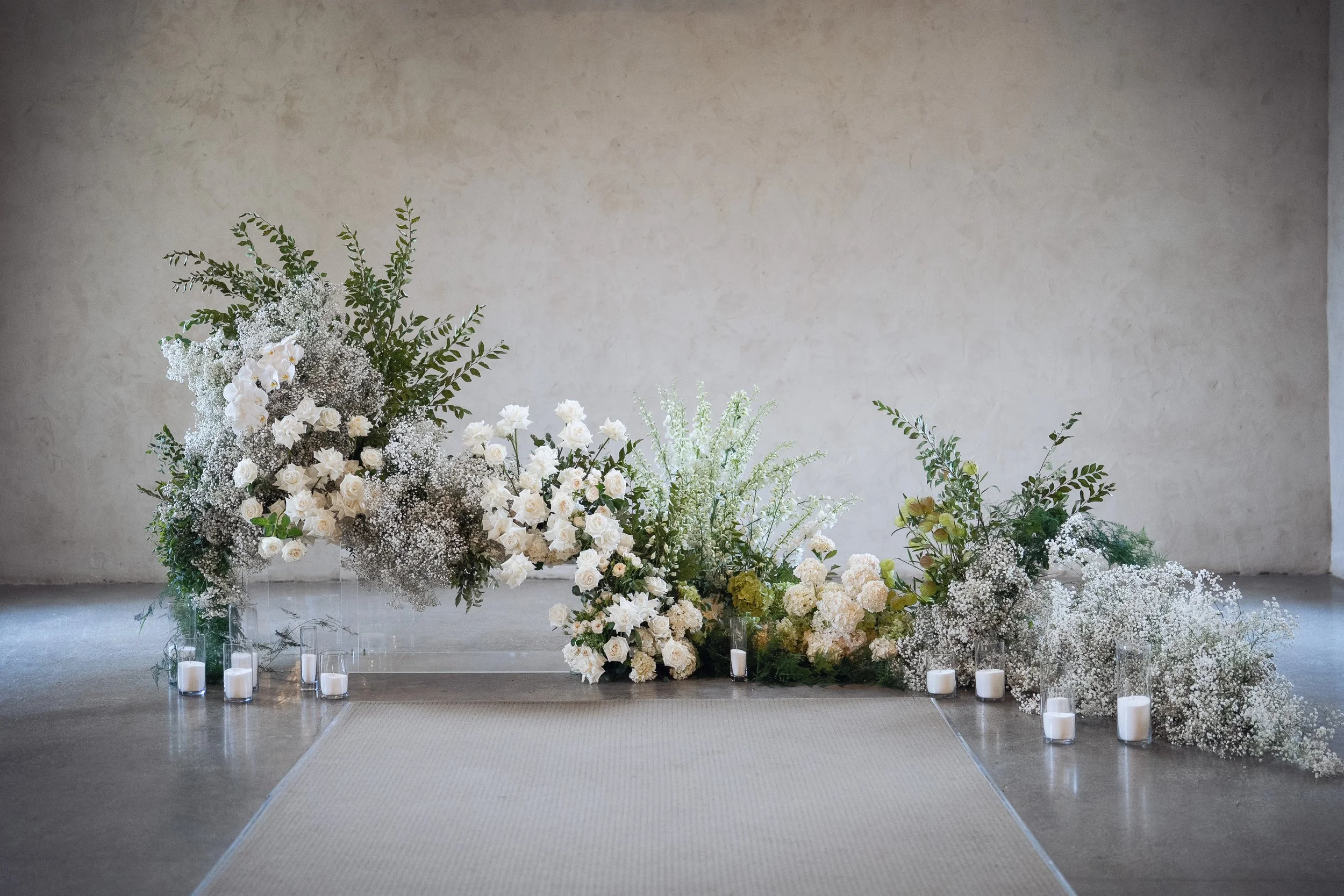 A floral arrangement with predominantly white flowers and greenery, set against a plain beige wall, with a few candles in glass holders placed on the floor nearby.