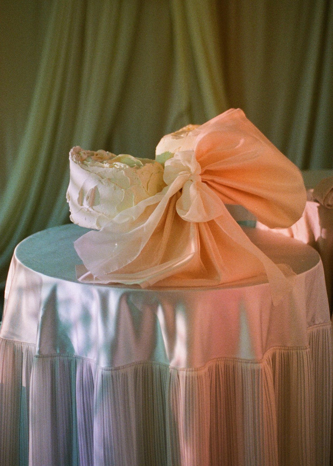 A wedding cake with pastel pink and white decorations, placed on a round table with a white tablecloth, against a background of cream-colored curtains.