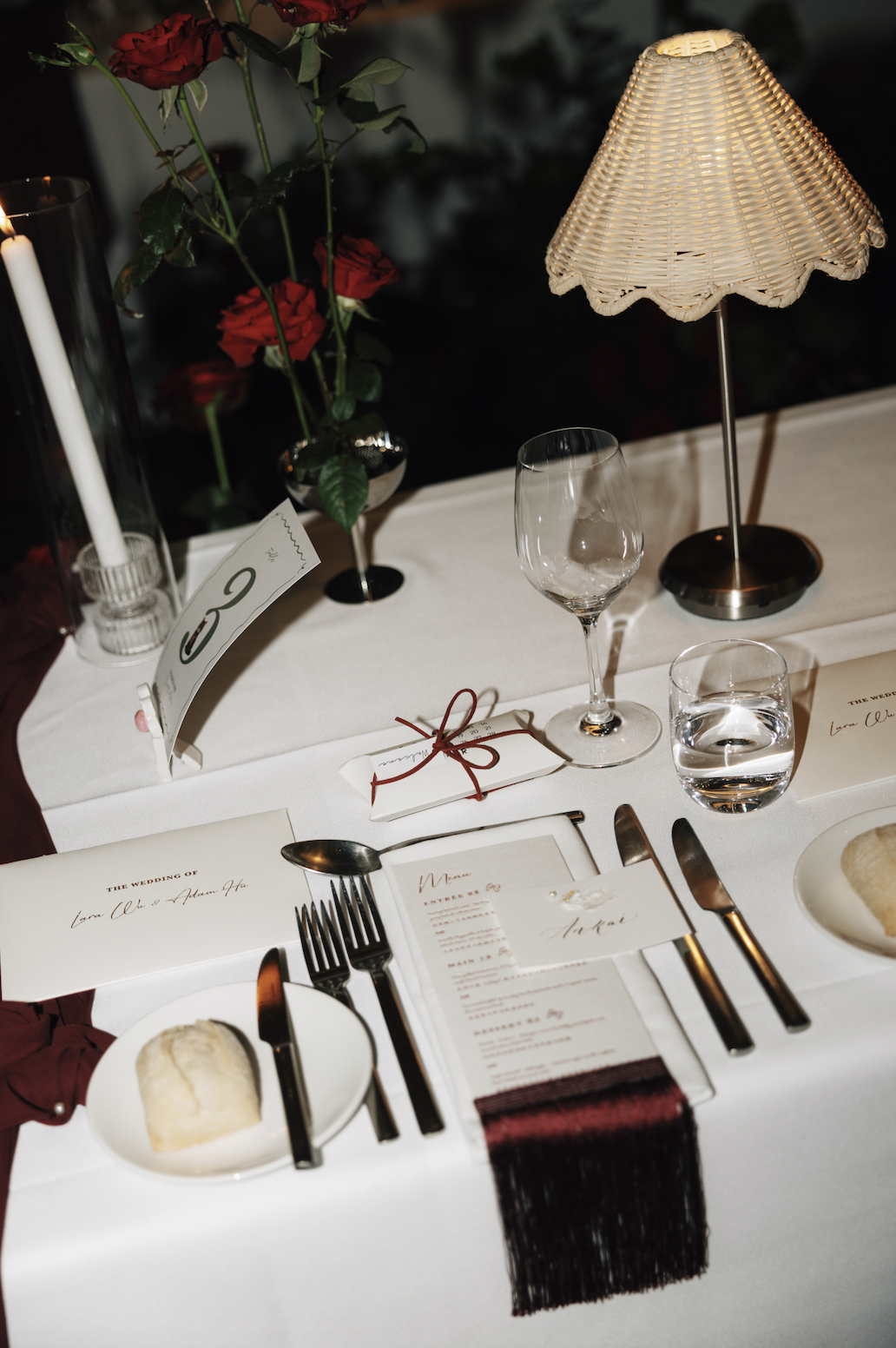 Elegant wedding reception table setting with white tablecloth, wine glasses, water glass, cutlery, a menu, a wrapped gift, roses in a vase, a lit candle, and a wicker lamp
