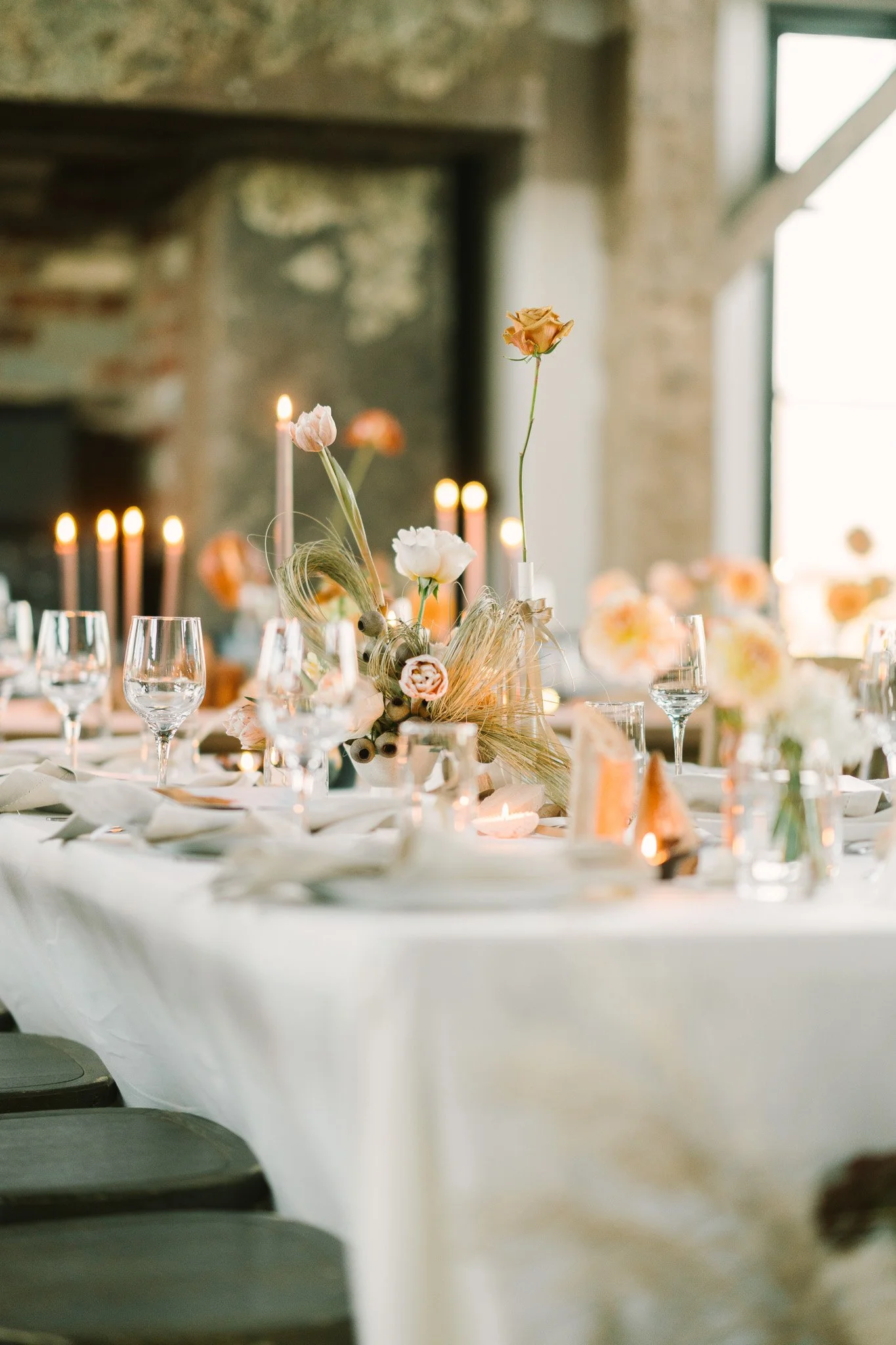 A decorated table with floral centerpieces, candles, wine glasses, and table settings in a rustic indoor space with stone walls and large windows.
