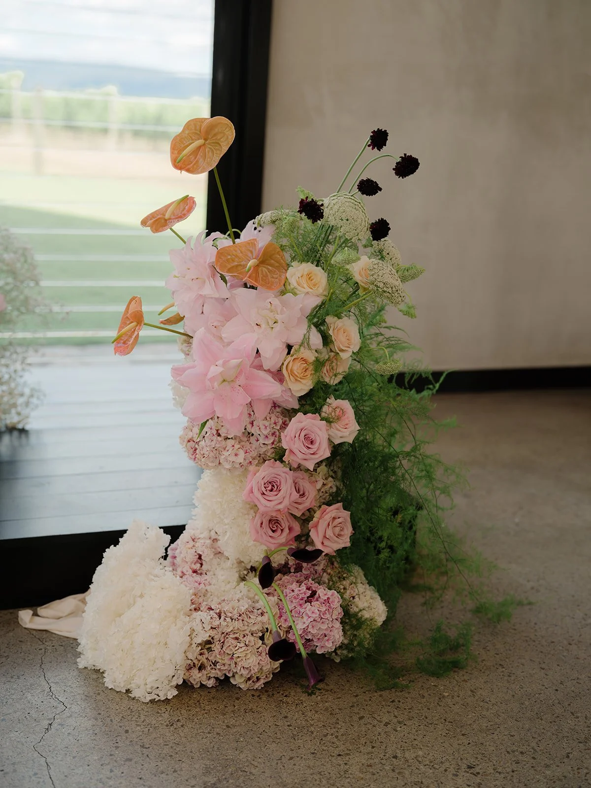 A flower arrangement with pink roses, white hydrangeas, and anthuriums, accented with dark purple thistles and greenery, placed on a concrete floor near a window.
