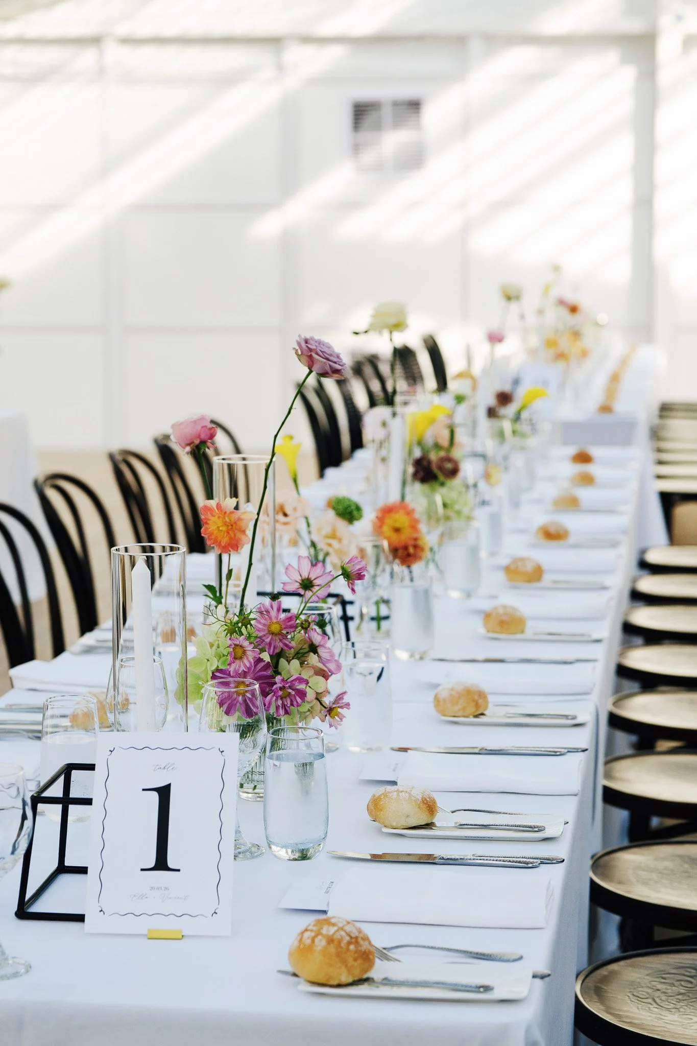 Long banquet table set with white tablecloths, decorated with colorful flowers in glass vases, and place settings including rolls, silverware, and water glasses. Black chairs line the sides of the table in a bright, airy room with high ceilings and l