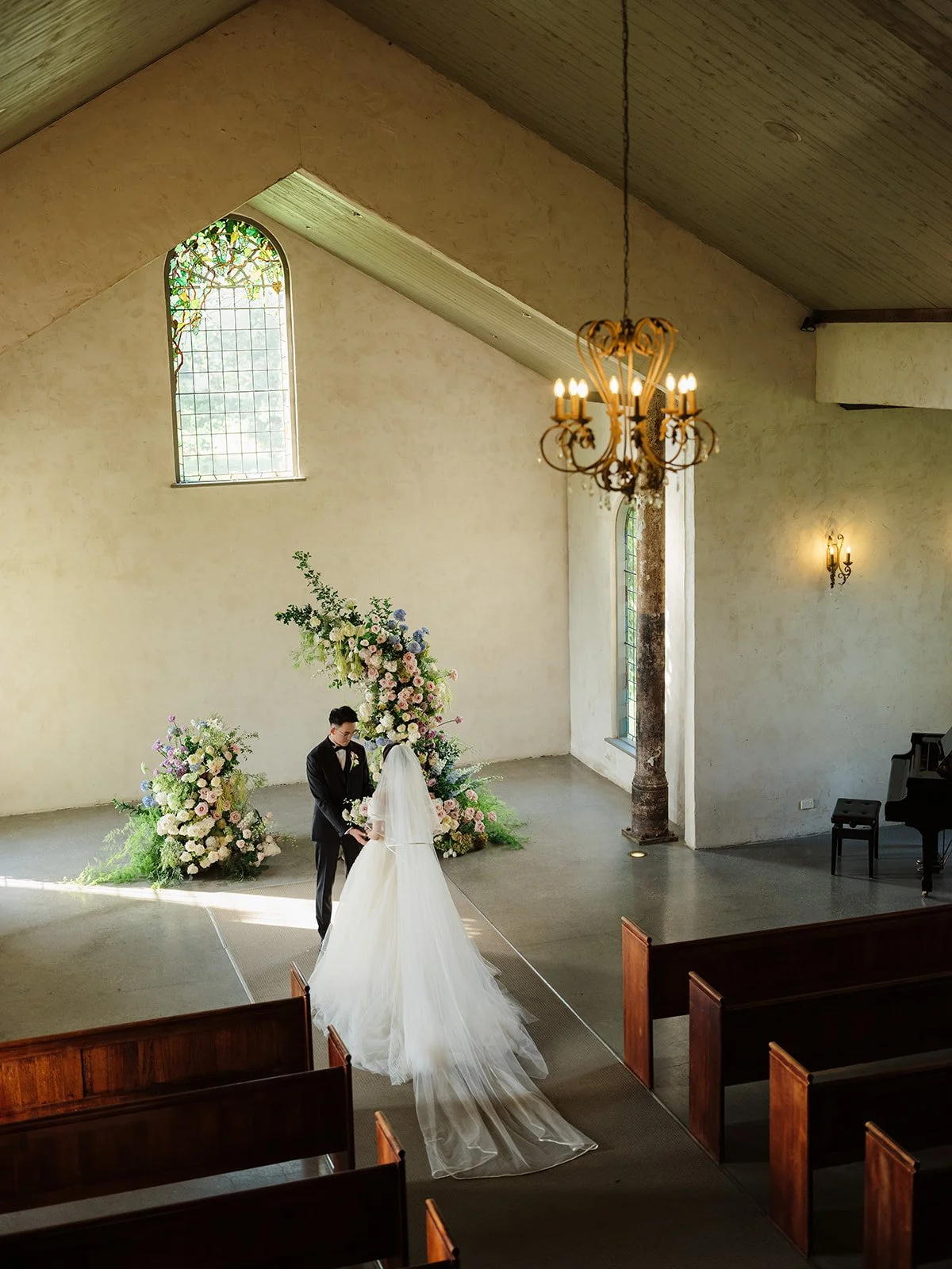 A bride and groom exchanging vows at an intimate wedding ceremony inside a church with high ceilings, stained glass windows, floral arrangements, a chandelier, and a piano.