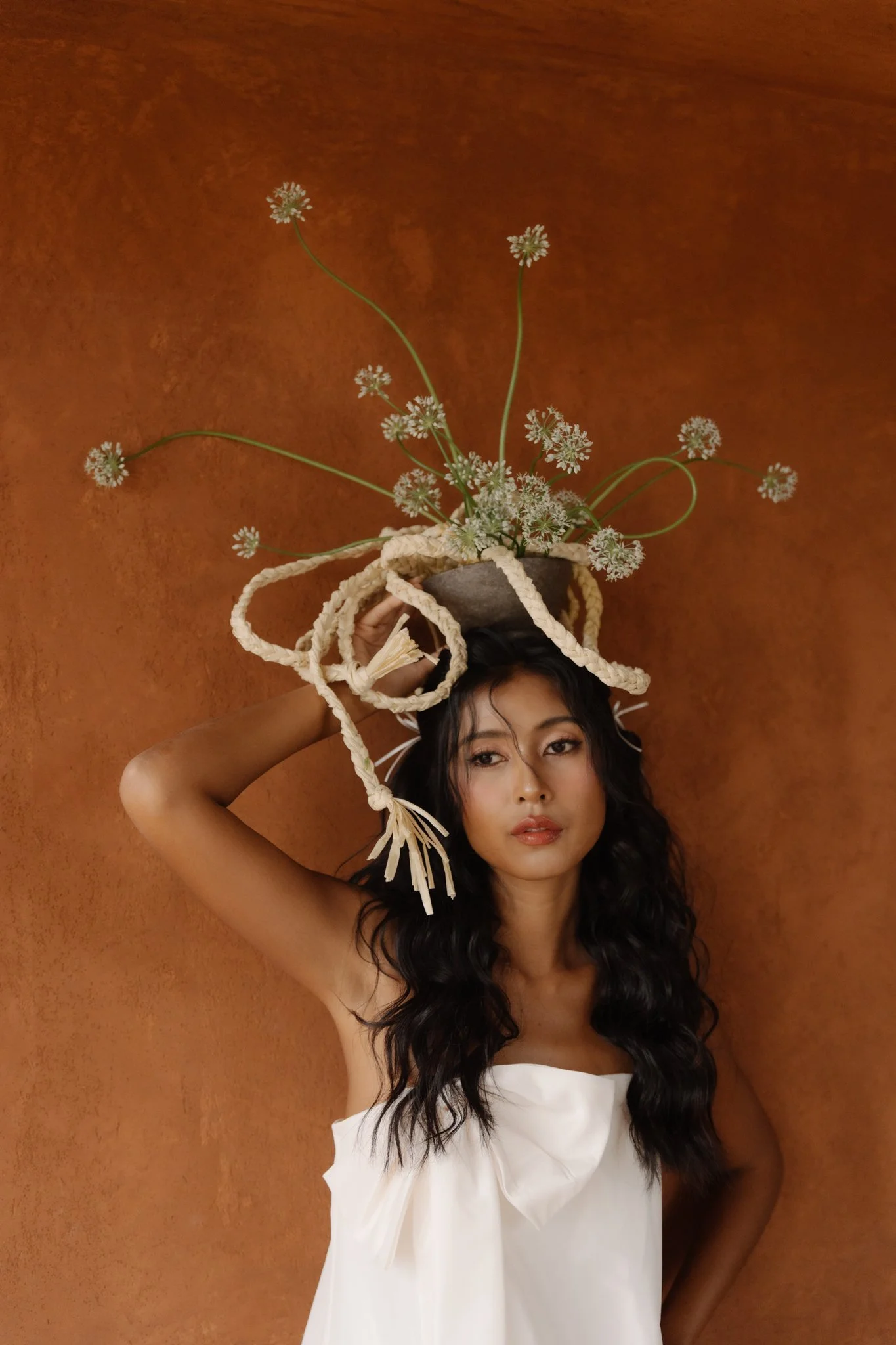 A woman with long dark wavy hair and light brown skin is holding a decorative arrangement of small white flowers in a dark gray pot above her head. She is wearing a white strapless dress and is standing against a rust-colored wall.