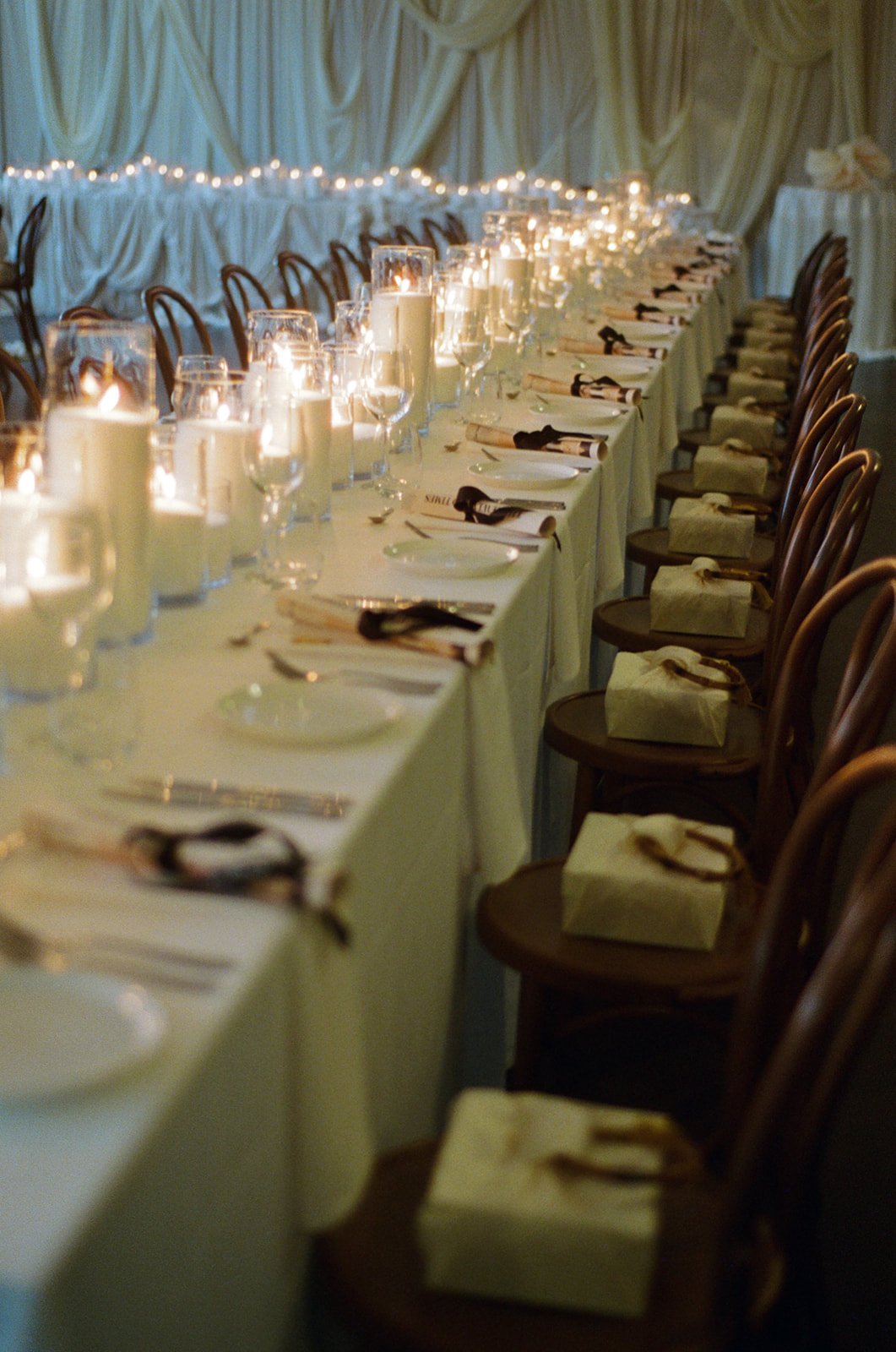 A long banquet table set with white tablecloth, candles in glass holders, wine glasses, plates, silverware, and wrapped napkins, in a decorated event hall with draped curtains and string lights.