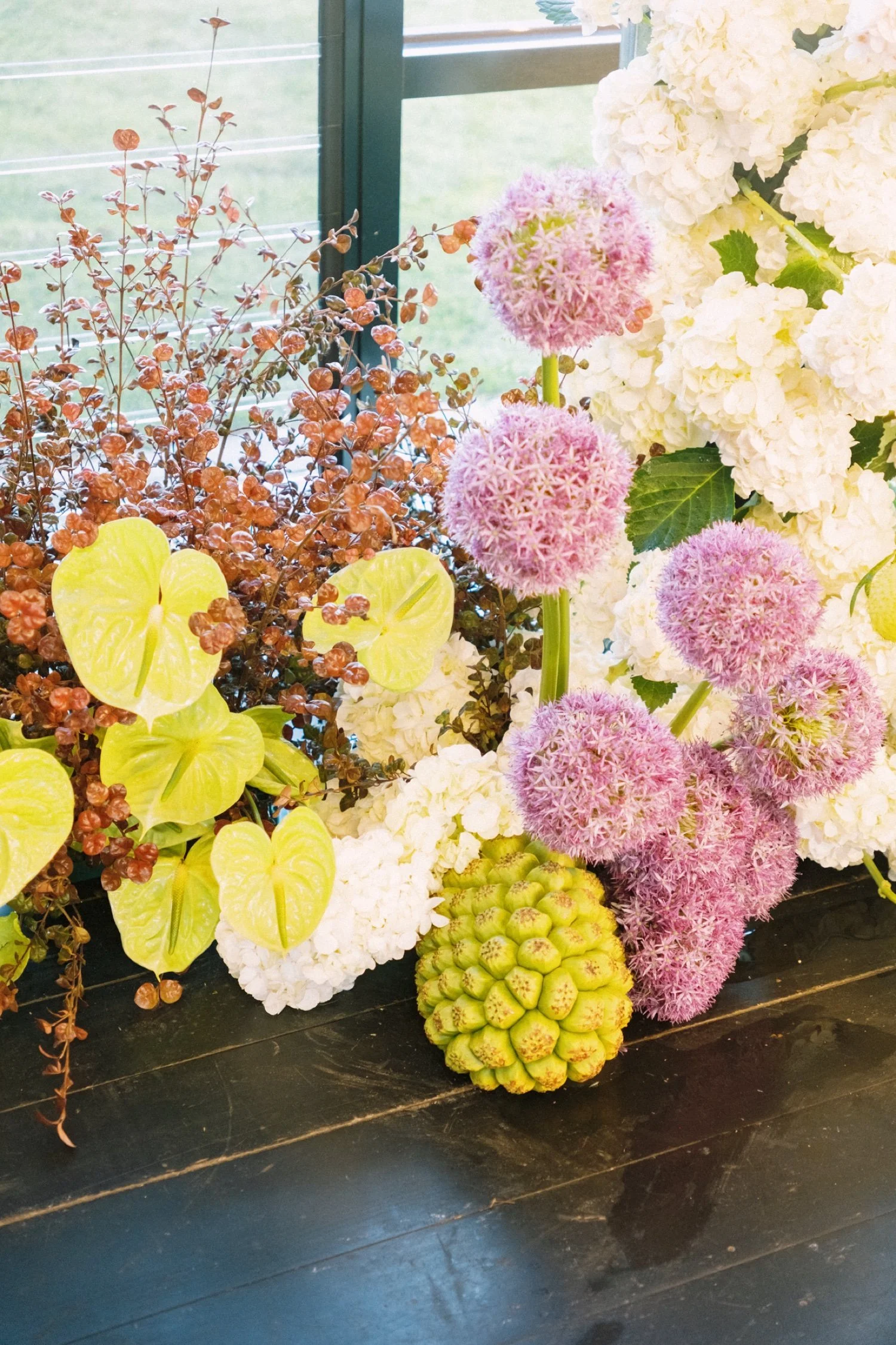 Colorful floral arrangement with pink spherical allium flowers, white hydrangeas, green pineapples, and yellow anthuriums on a dark wooden surface, with a window and green lawn in the background.