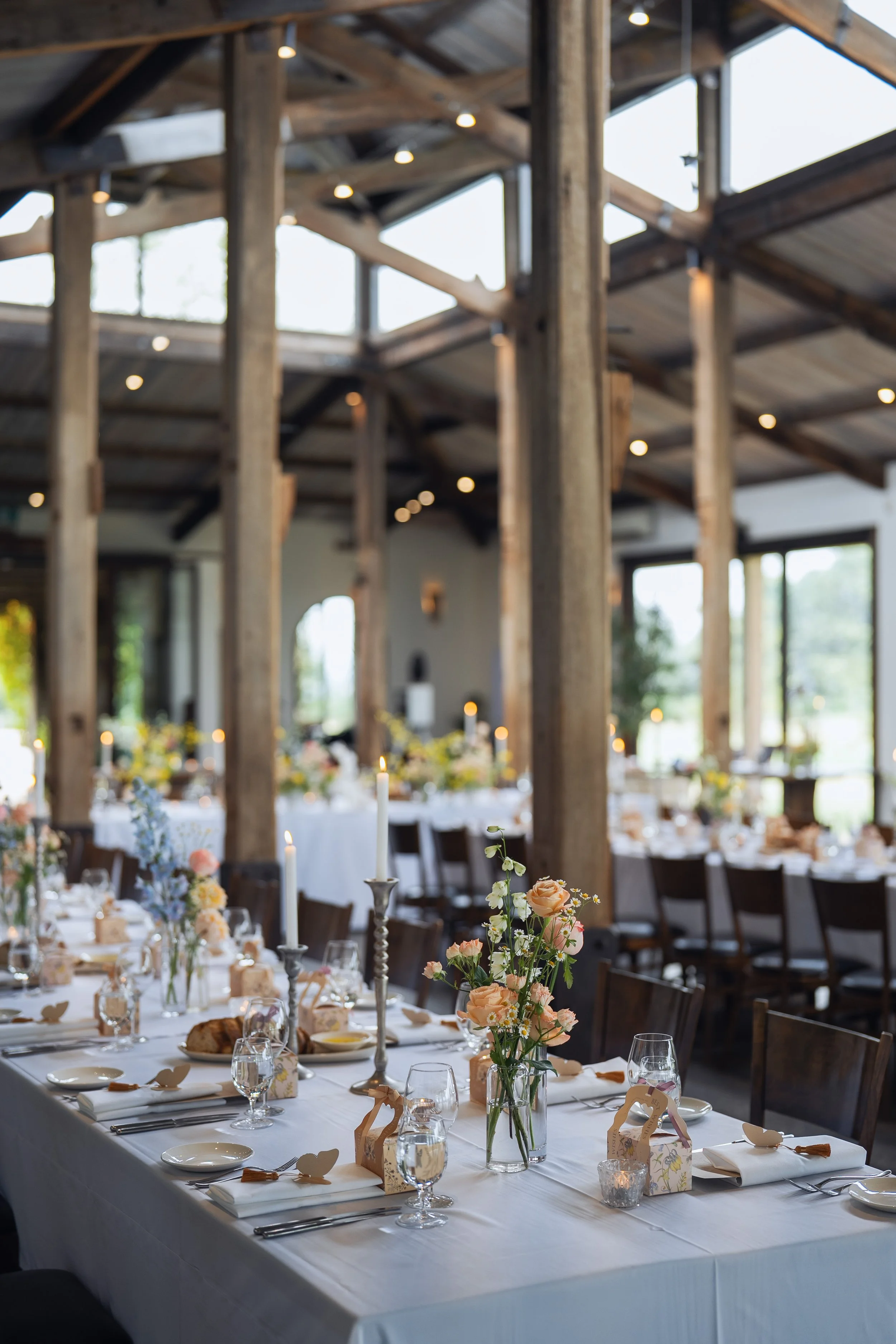 Indoor wedding reception with long rectangular table, floral centerpieces, candles, and place settings, in a rustic venue with wooden beams and large windows.