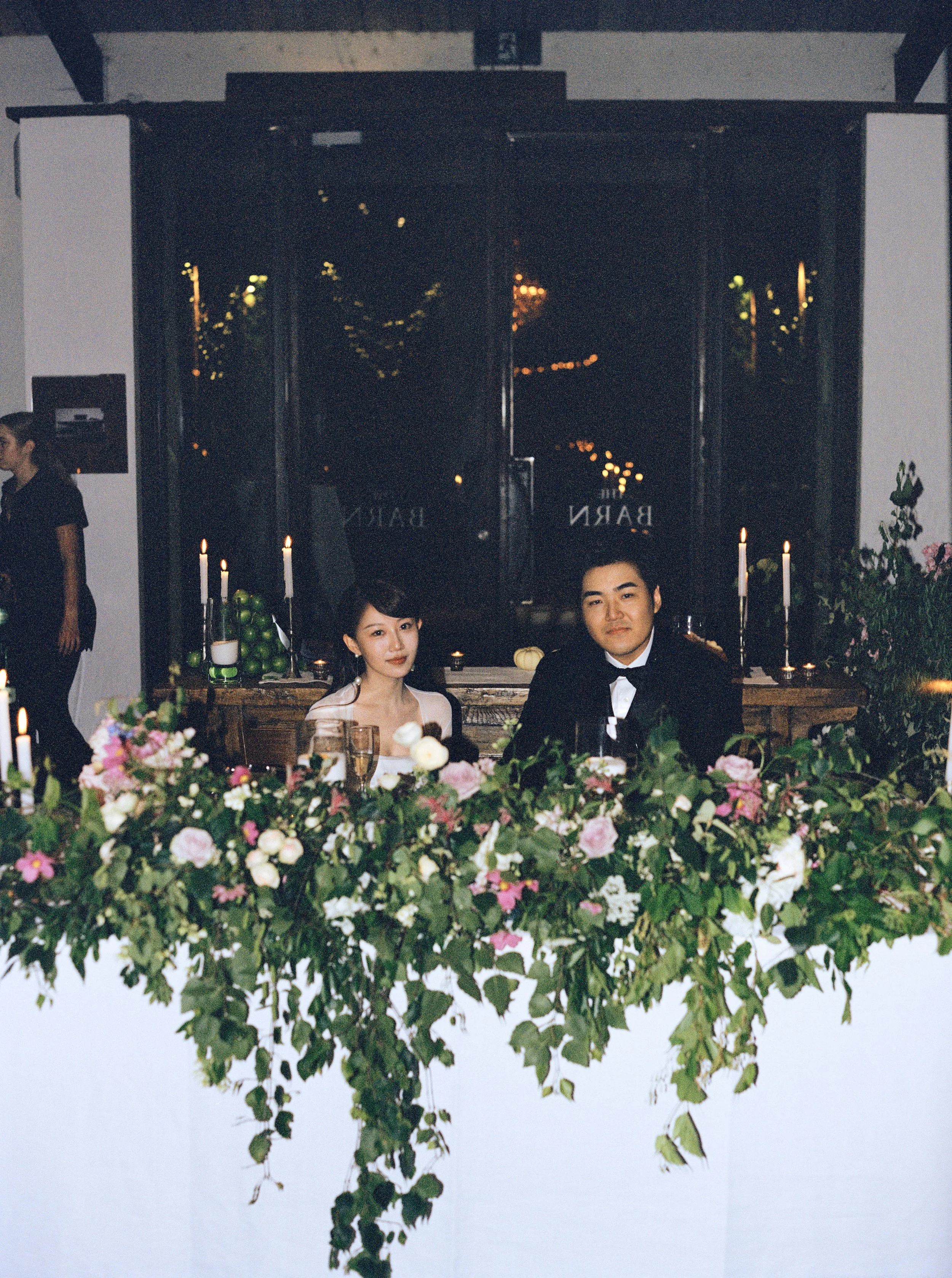 People sitting at a table decorated with flowers and candles, with a floral wall and large reflective window in the background.