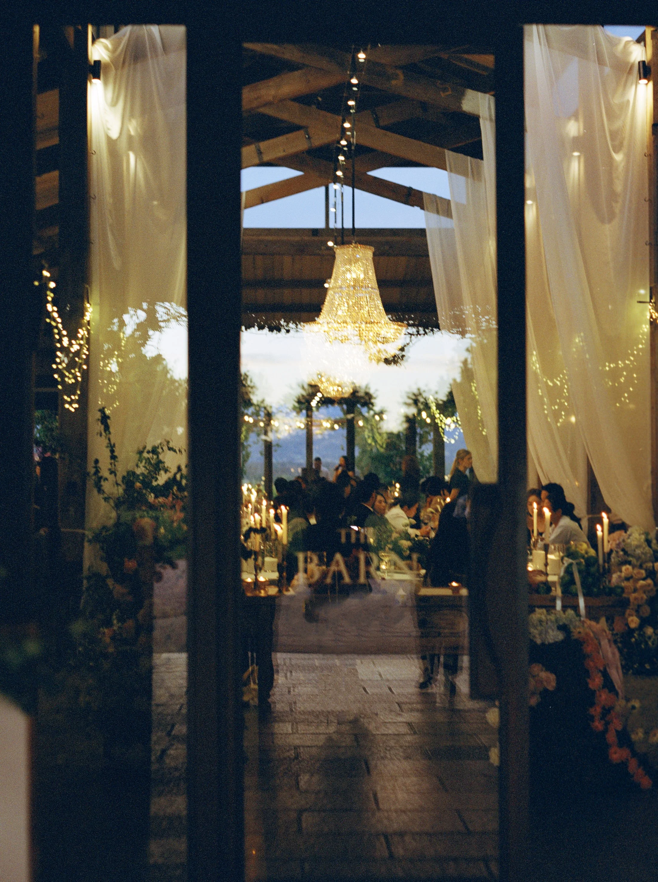 A view through a dark doorway into a decorated wedding reception area with warm lighting, candles, flowers, and a chandelier hanging from a wooden ceiling.