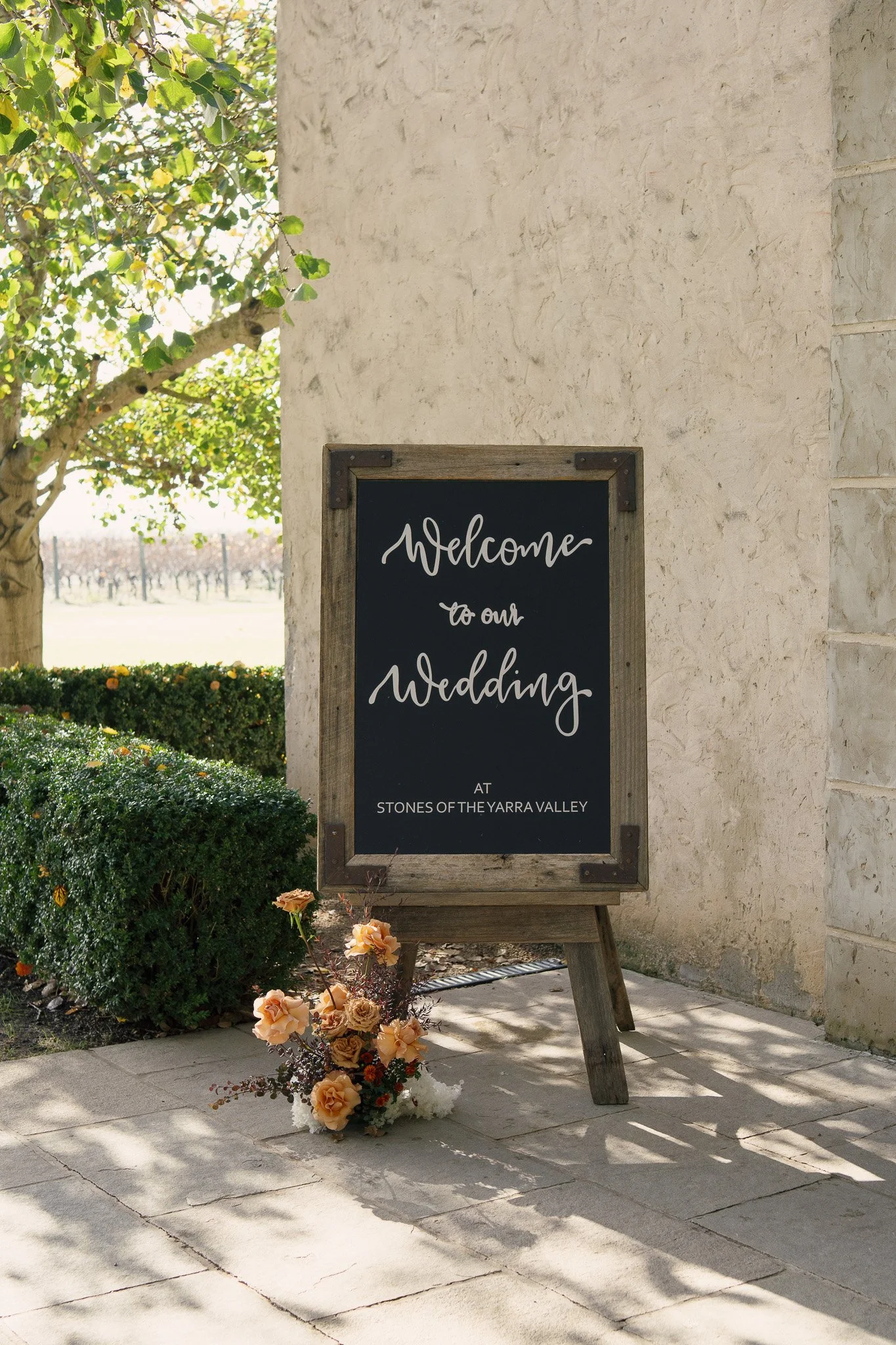 Outdoor wedding reception signboard with floral decoration in a landscaped garden, near a stone wall, with a tree in the background.