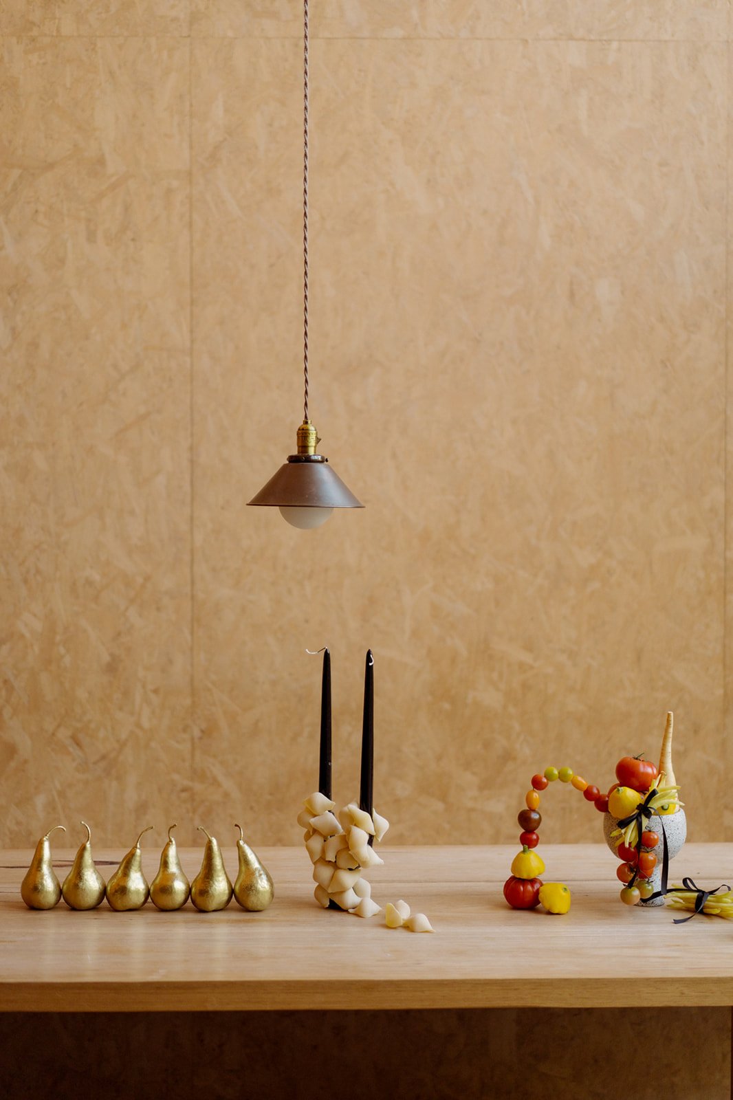 A minimalistic arrangement of objects on a wooden surface against a beige wall. Features a vintage pendant lamp, six gold-colored pears, black taper candles with garlic cloves, and a vase with fresh tomatoes, garlic, and peppers.