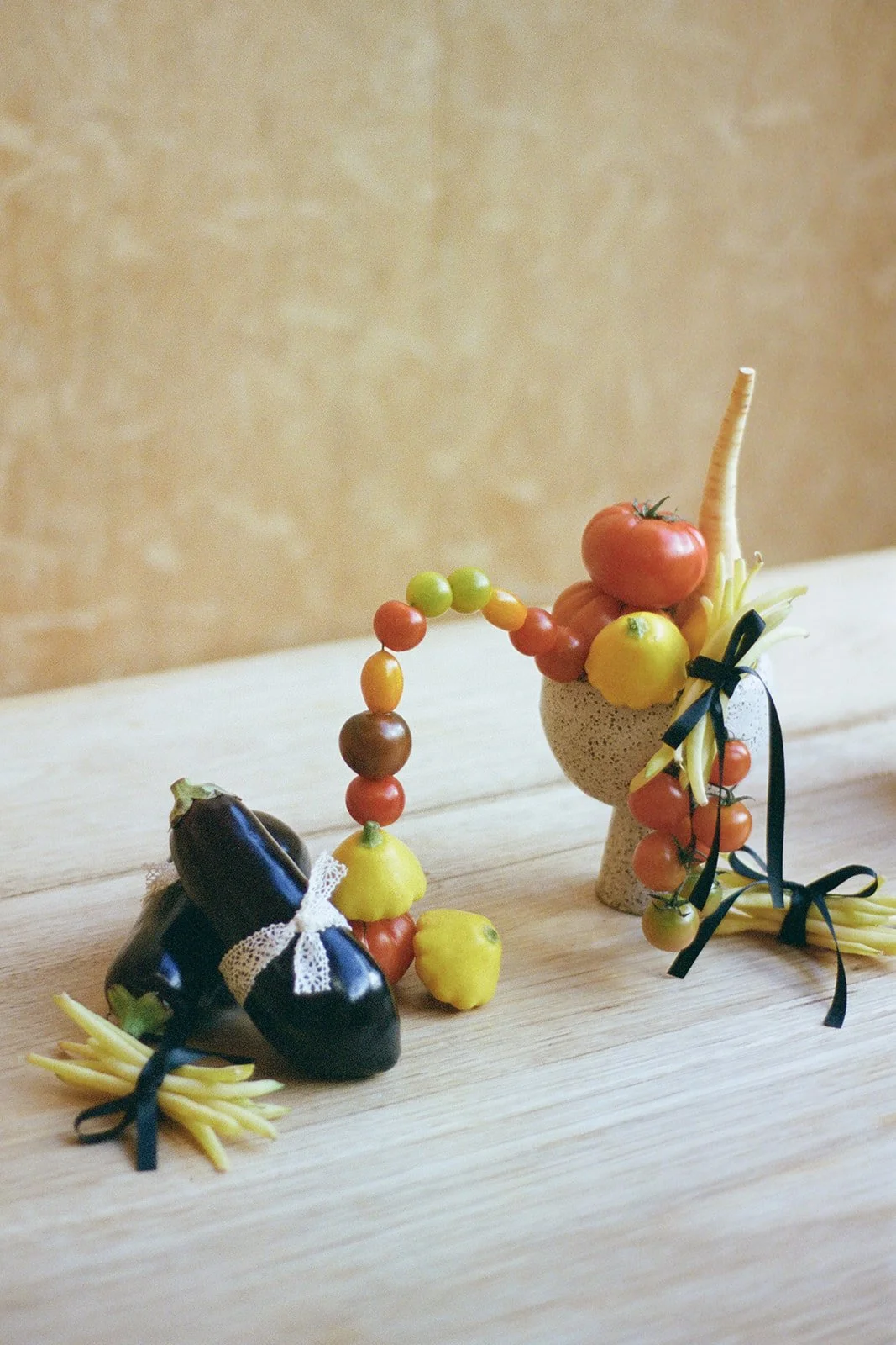 A collection of small vegetables and fruits arranged on a wooden surface, including cherry tomatoes, grapes, an eggplant, a lemon, a yellow squash, a carrot, and green onions, decorated with black ribbons and lace.