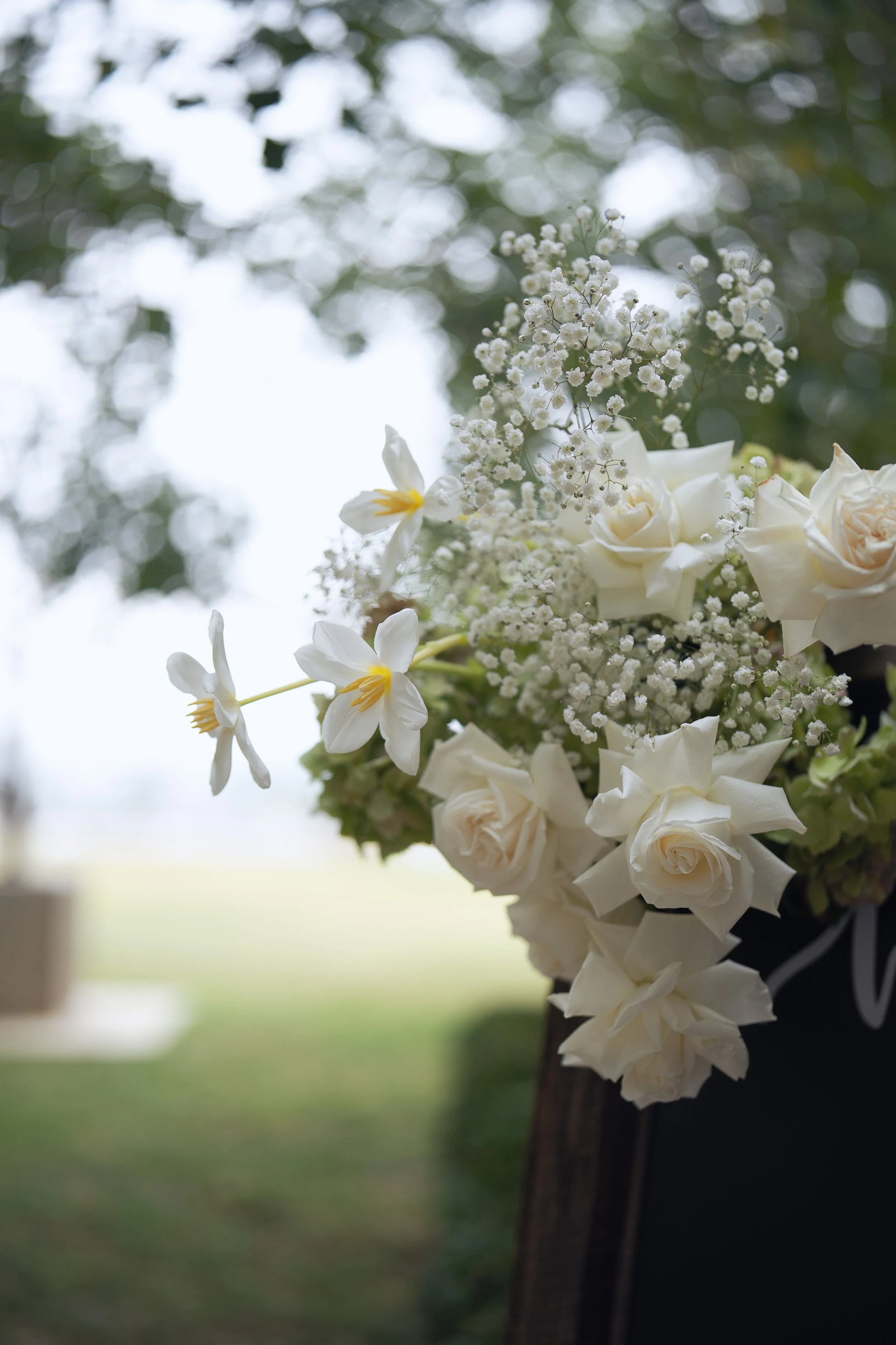 Close-up of a bouquet of white flowers, including roses and baby's breath, attached to a wooden post outdoors with a blurred background of greenery.