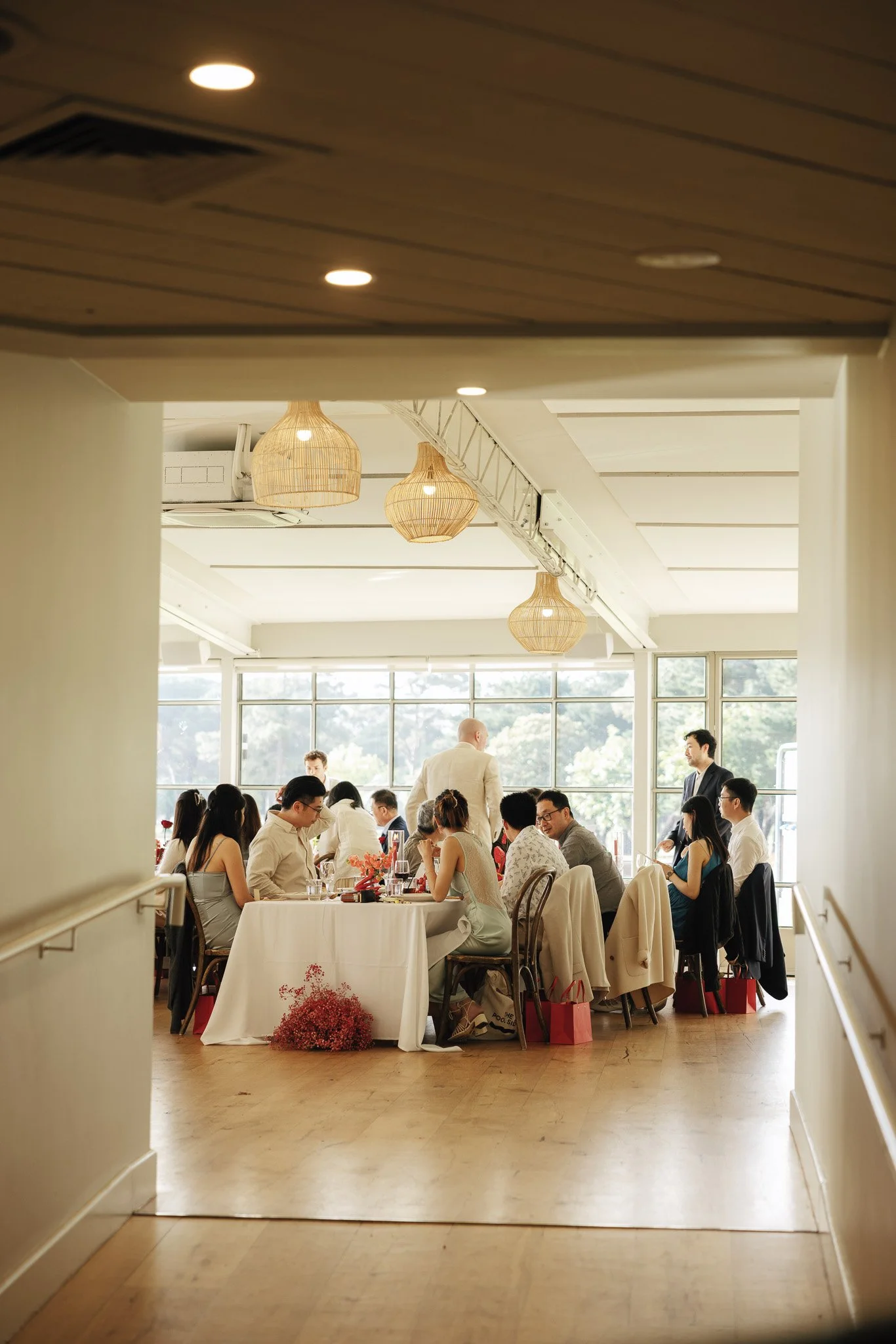 People gathered around tables at a banquet-style event in a well-lit, modern room with large windows and hanging wicker lamps.