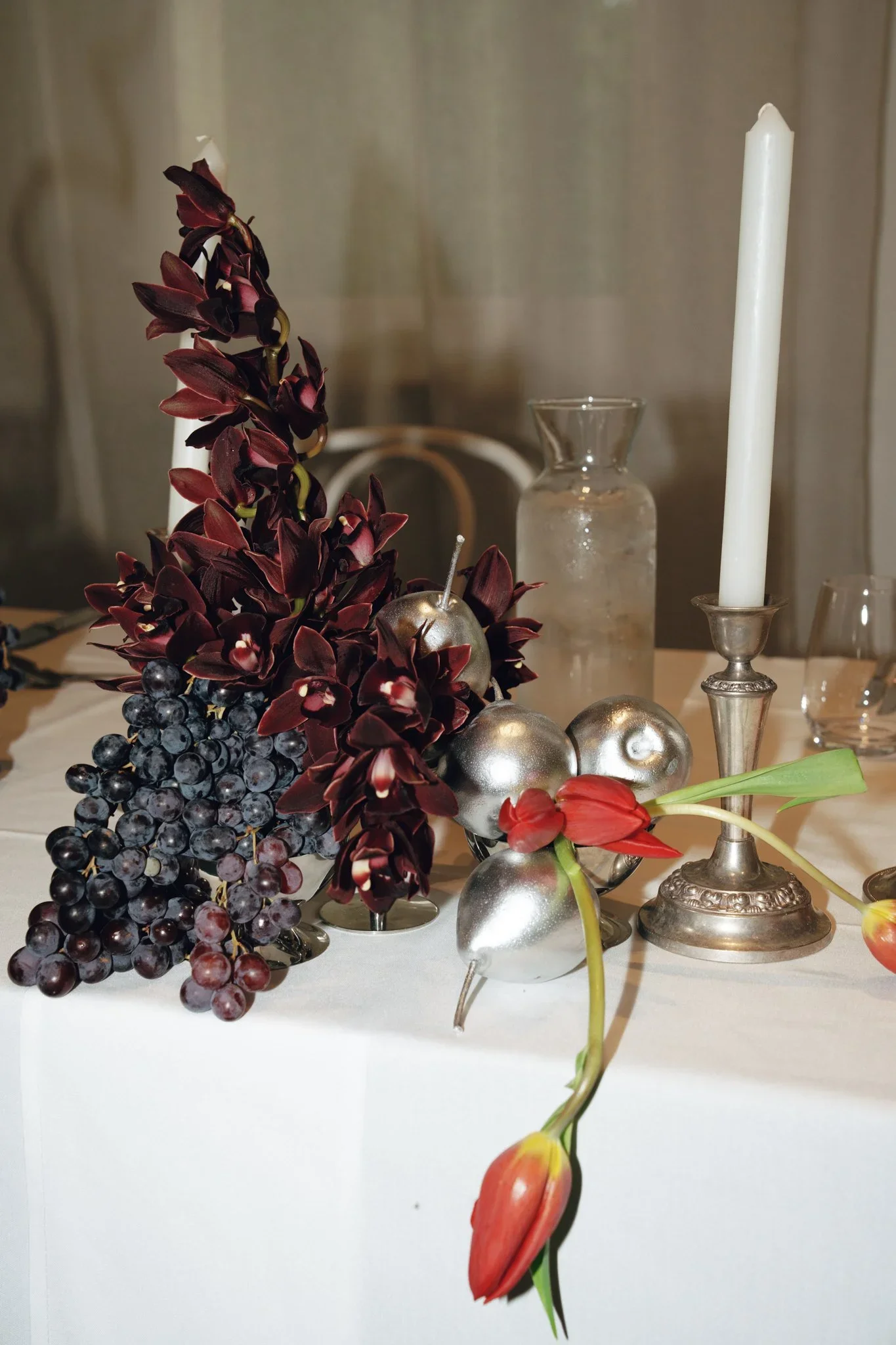 Decorative arrangement of dark red orchids, purple grapes, and silver ornaments on a white surface, with tall white candles and glass vases in the background.