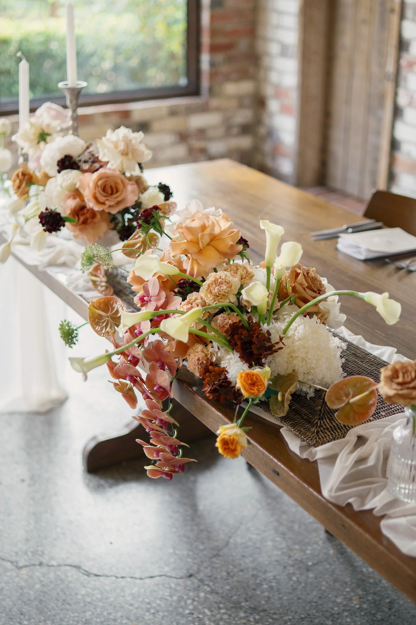 A long wooden table decorated with a floral arrangement of peach, white, and burgundy flowers, with candles and tableware set for a special occasion in a rustic room with a window and brick walls.
