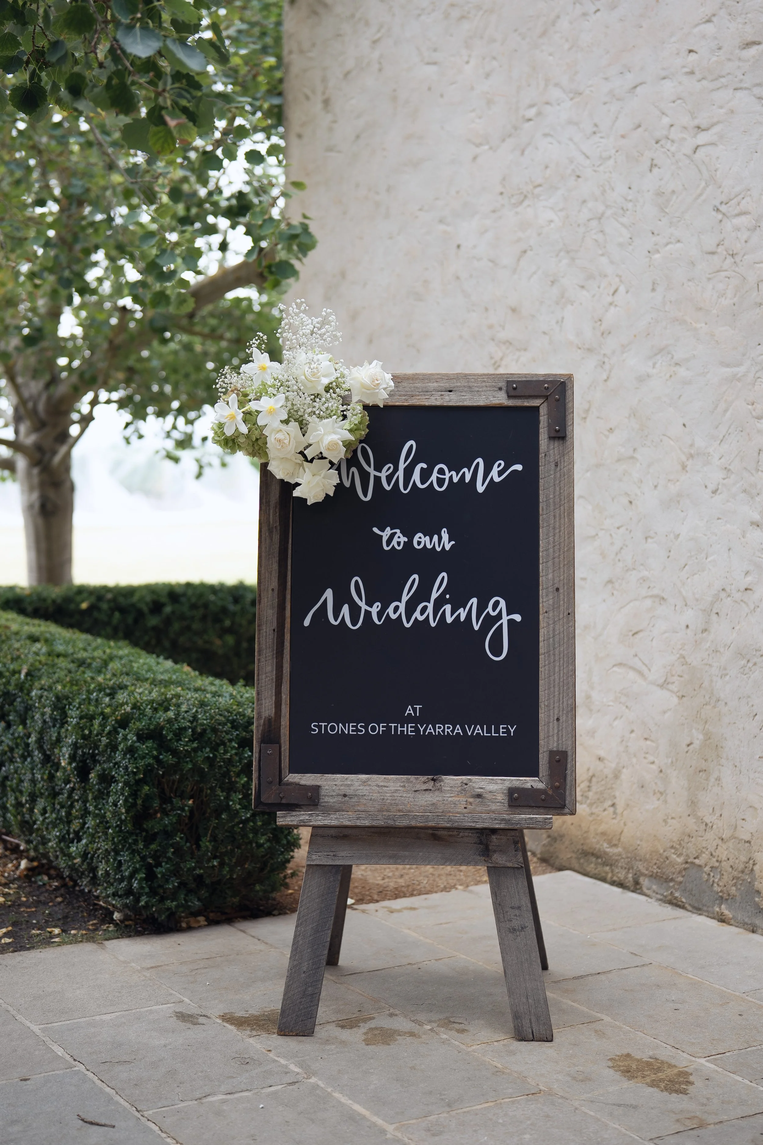 A chalkboard sign decorated with white flowers on a wooden frame, placed outdoors on a sidewalk, welcoming guests to a wedding at Stones of the Yarra Valley, next to a cream-colored textured wall and some greenery.