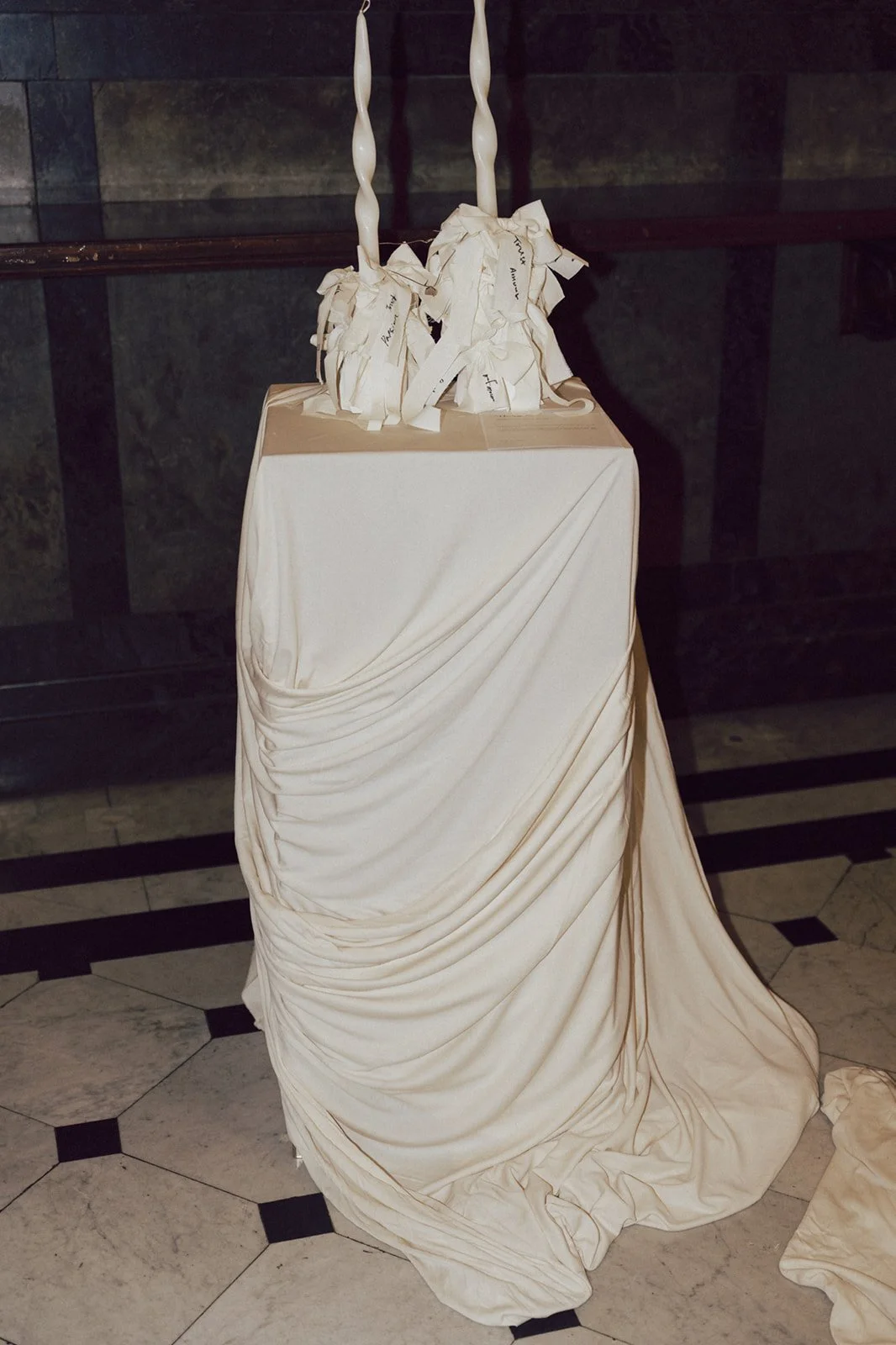 A white cloth draped over a box with gathered fabric and ruched details, topped with an artistic white sculpture with two twisted, candle-like elements, set against a dark wall.