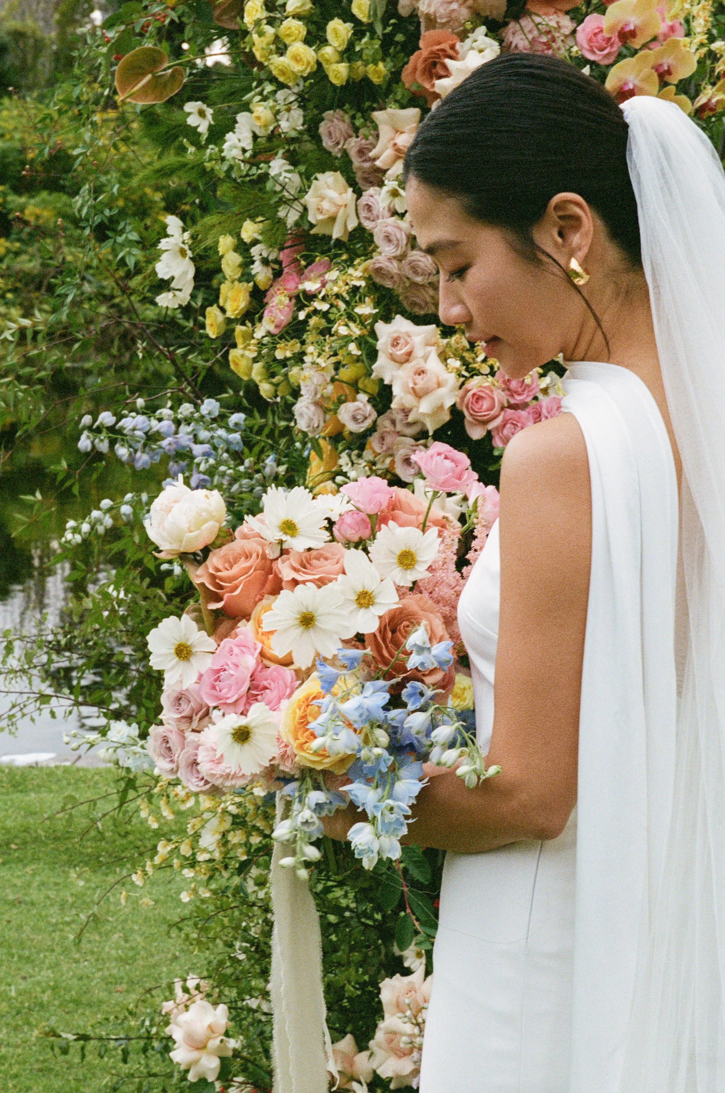 A bride in a white wedding dress and veil stands beside a large floral decoration made of pink, white, yellow, and purple flowers, with green foliage in the background.