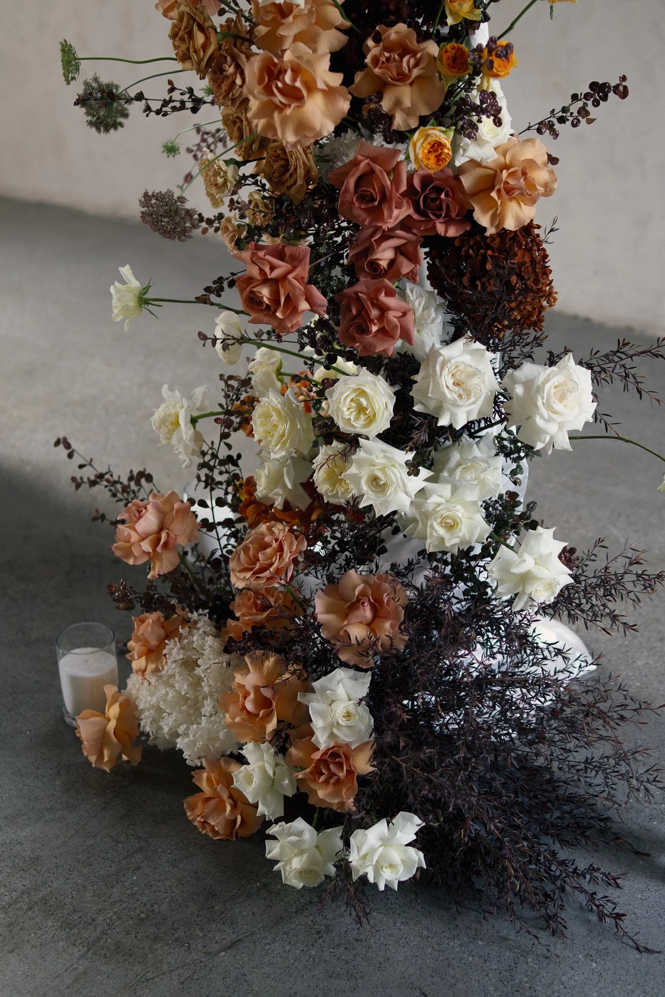 A tall floral arrangement of cream, peach, and rust-colored roses, white hydrangeas, purple and green accents, and dark burgundy foliage, beside a lit white candle on a concrete surface.
