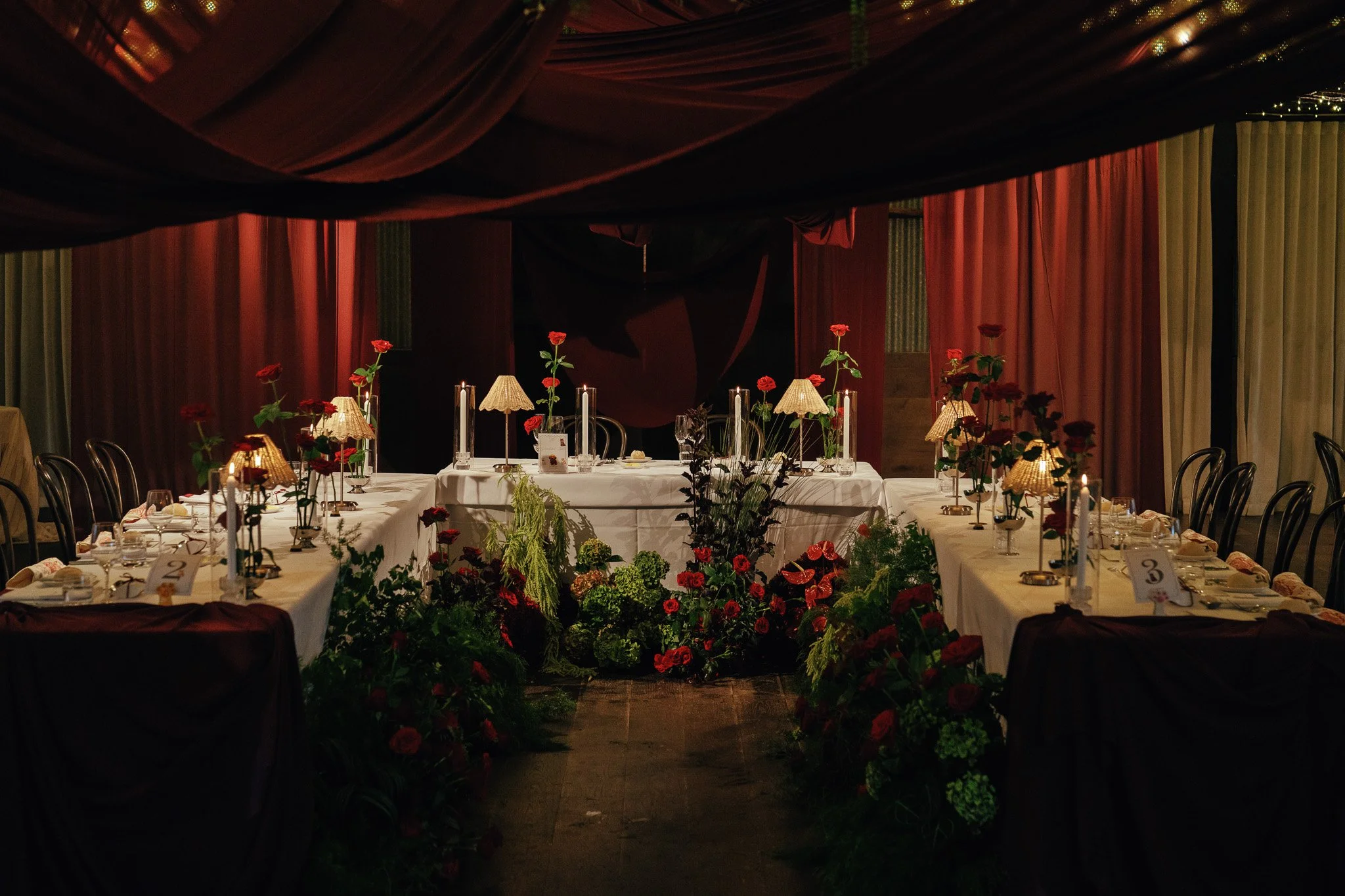 Decorated banquet hall with tables, red roses, small lamps, and red velvet drapes.