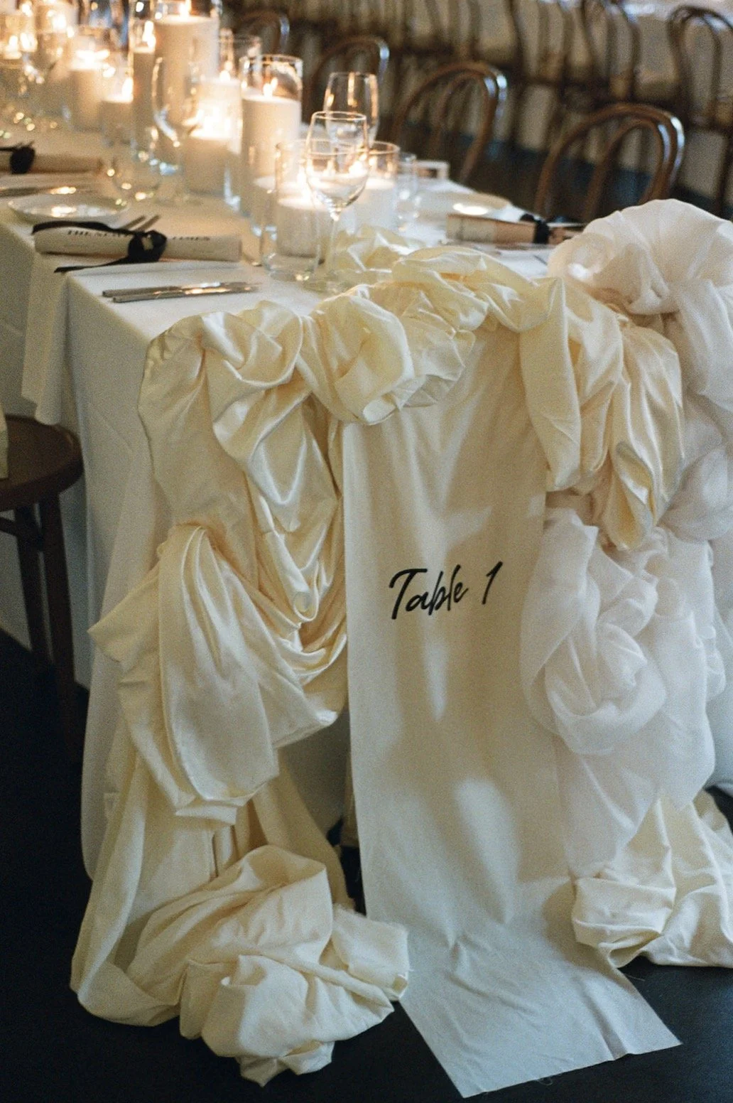 A decorated banquet table with candles and glassware, with a chair draped in a cream-colored fabric that has a ruffled design, and a sign that says 'Table 1' attached to the fabric.