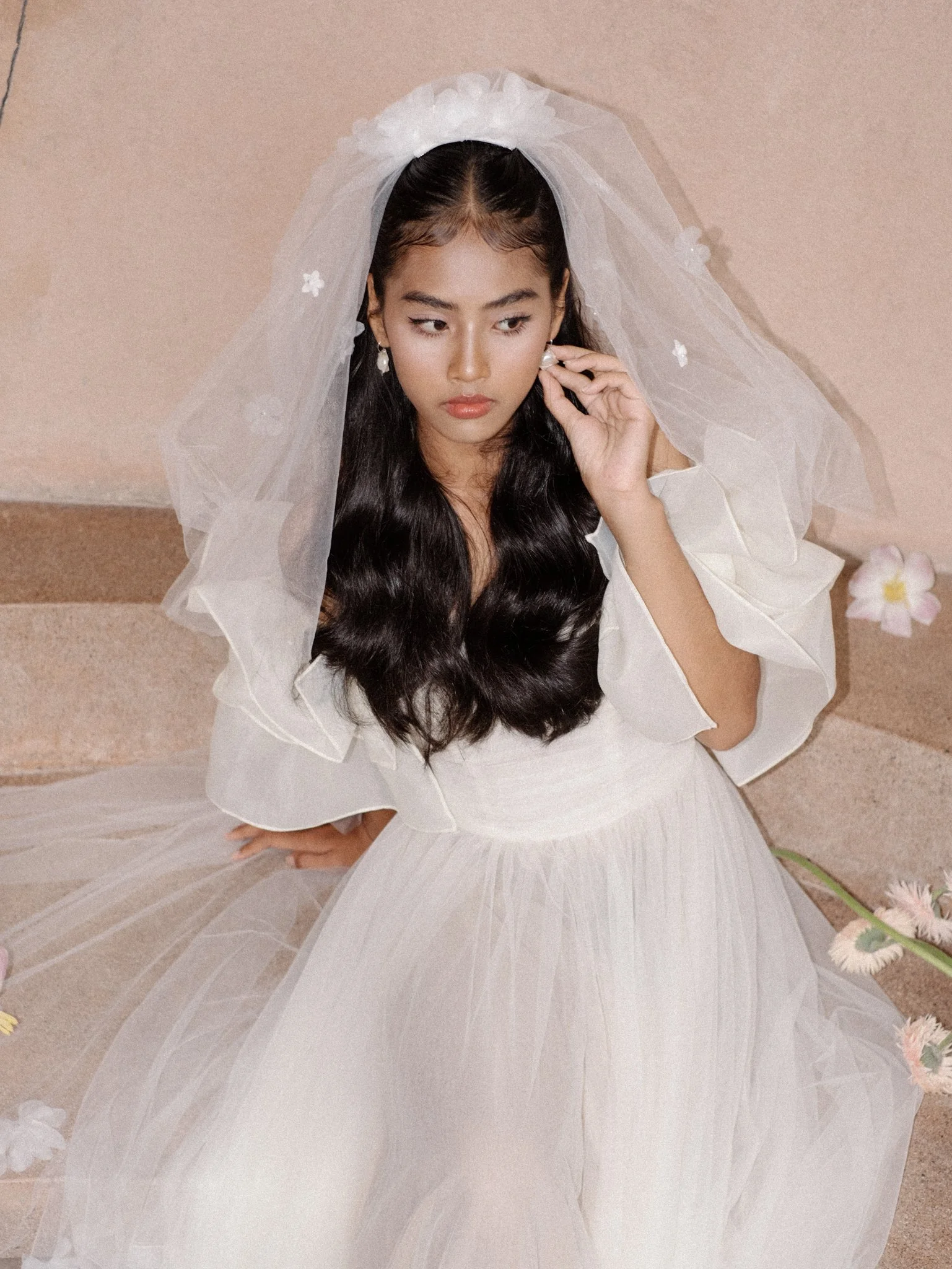 A young woman dressed in a white gown and veil, likely a bride, posed indoors on beige stairs with floral decorations around, looking to the side with a contemplative expression.