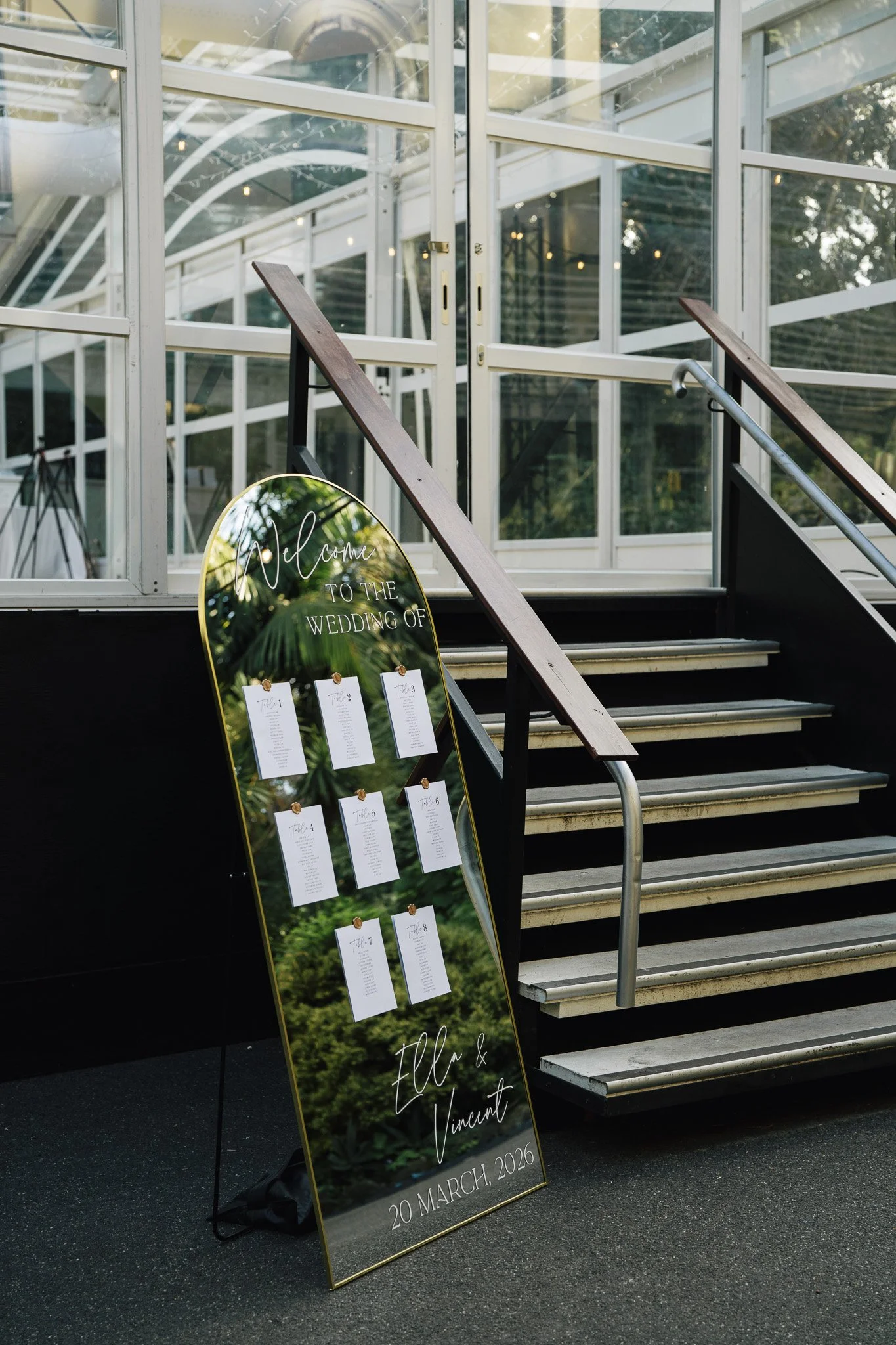 A large wedding sign with a forest background, featuring table numbers and the names Ellie and Vincent, with the wedding date March 20, 2026, placed at the bottom of a set of stairs outside a glass building.