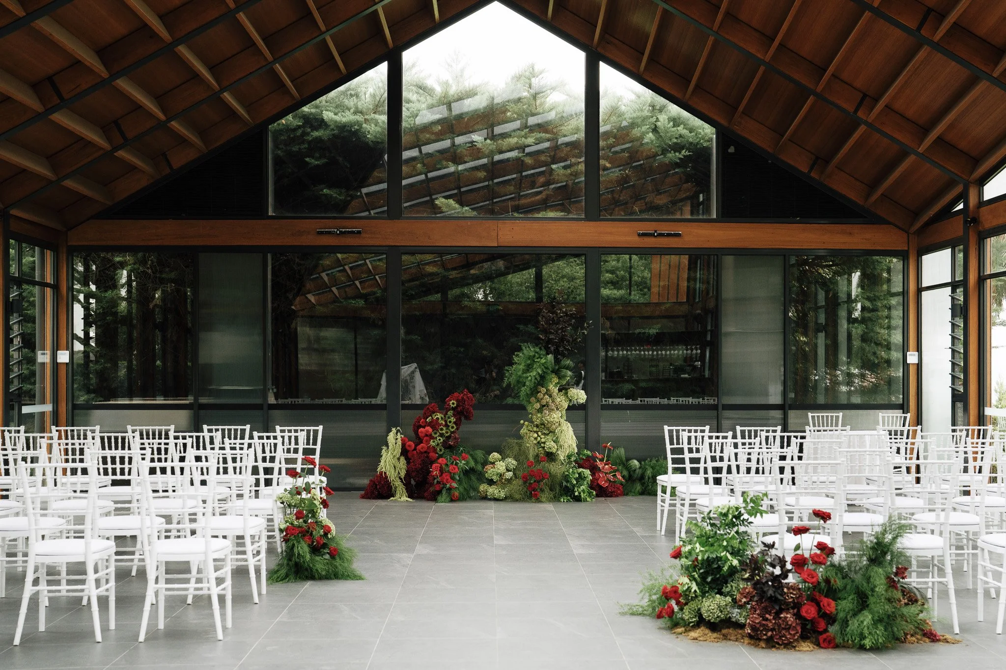 Indoor event space with white chairs arranged in rows, decorated with red and green floral arrangements, set in front of large glass windows and a wooden ceiling.