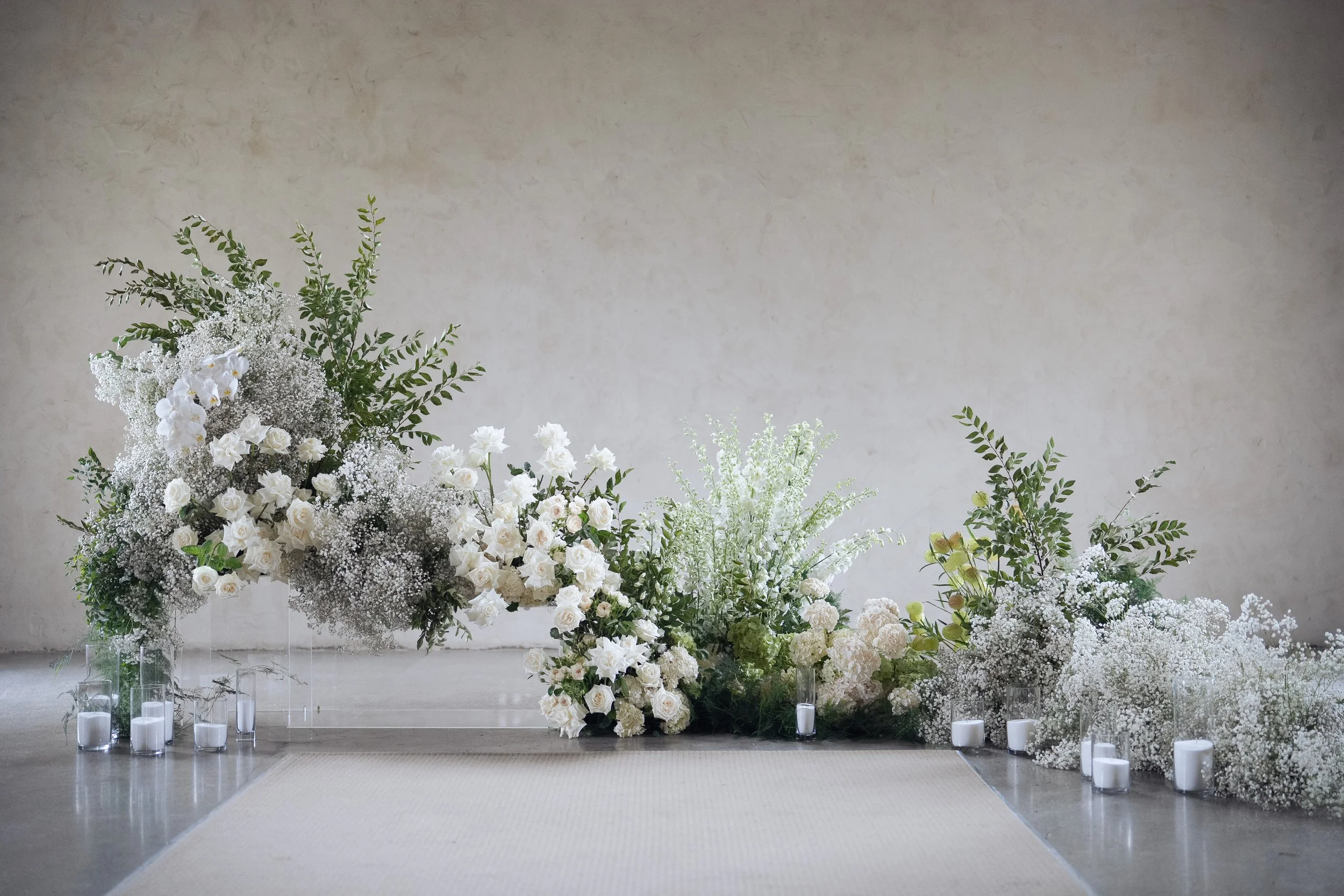 Arrangement of white flowers and greenery with candles on the floor, set against a plain wall, likely for a wedding or special event.