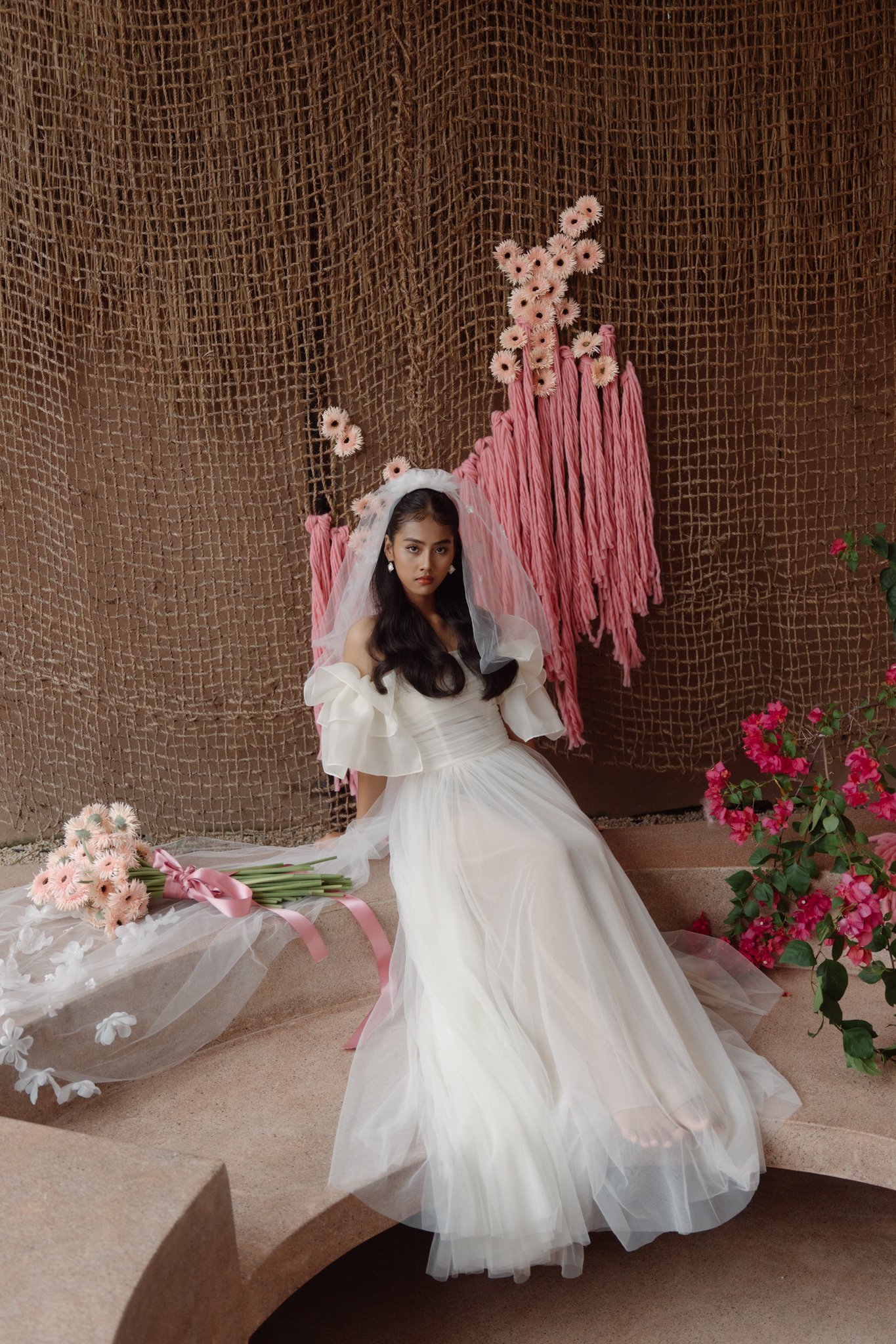 A woman dressed in a white gown with puffed sleeves and a tulle veil sitting on steps in front of a brown textured backdrop with pink flowers and flowers lying on the ground.