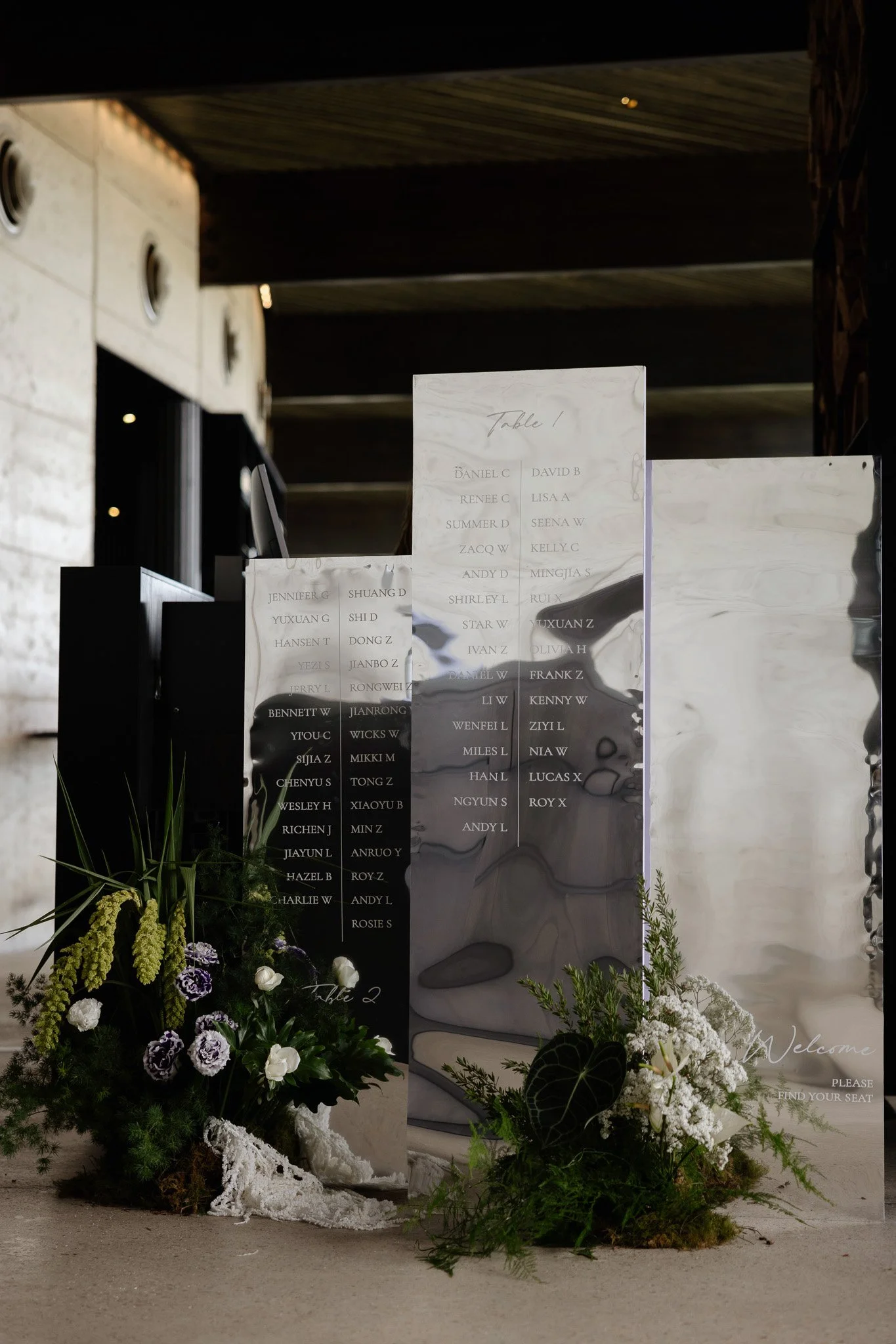 Wedding seating display with floral arrangements and black and white seating charts. The image features a floral arrangement with purple, white, and green flowers at the bottom left. The seating charts are hanging vertically, with the one on the righ