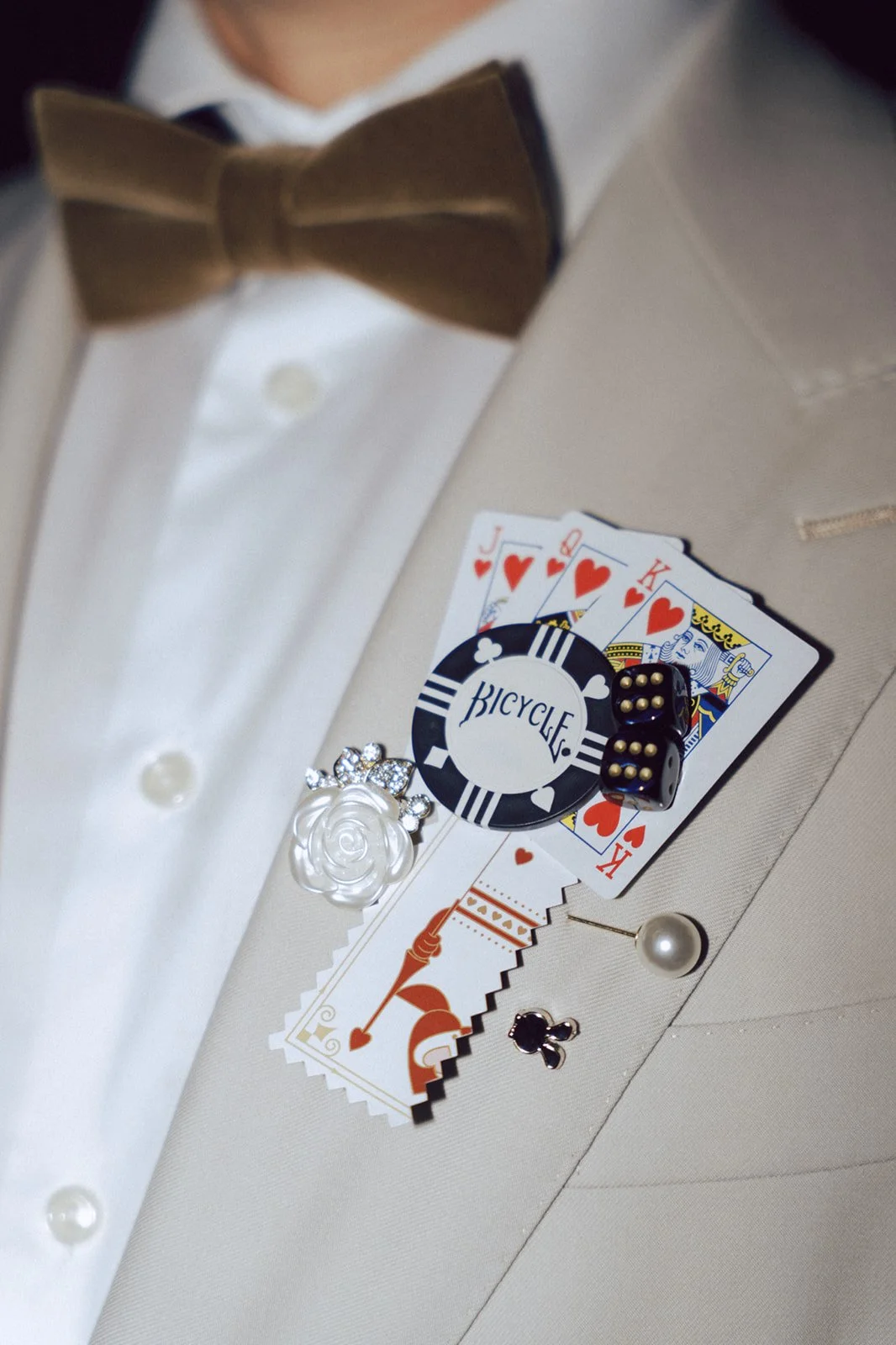 A collection of playing cards, a bicycle poker chip, two black dice with gold dots, a pearl earrings, and a white flower pin on a light beige suit jacket with a white shirt and brown bow tie.