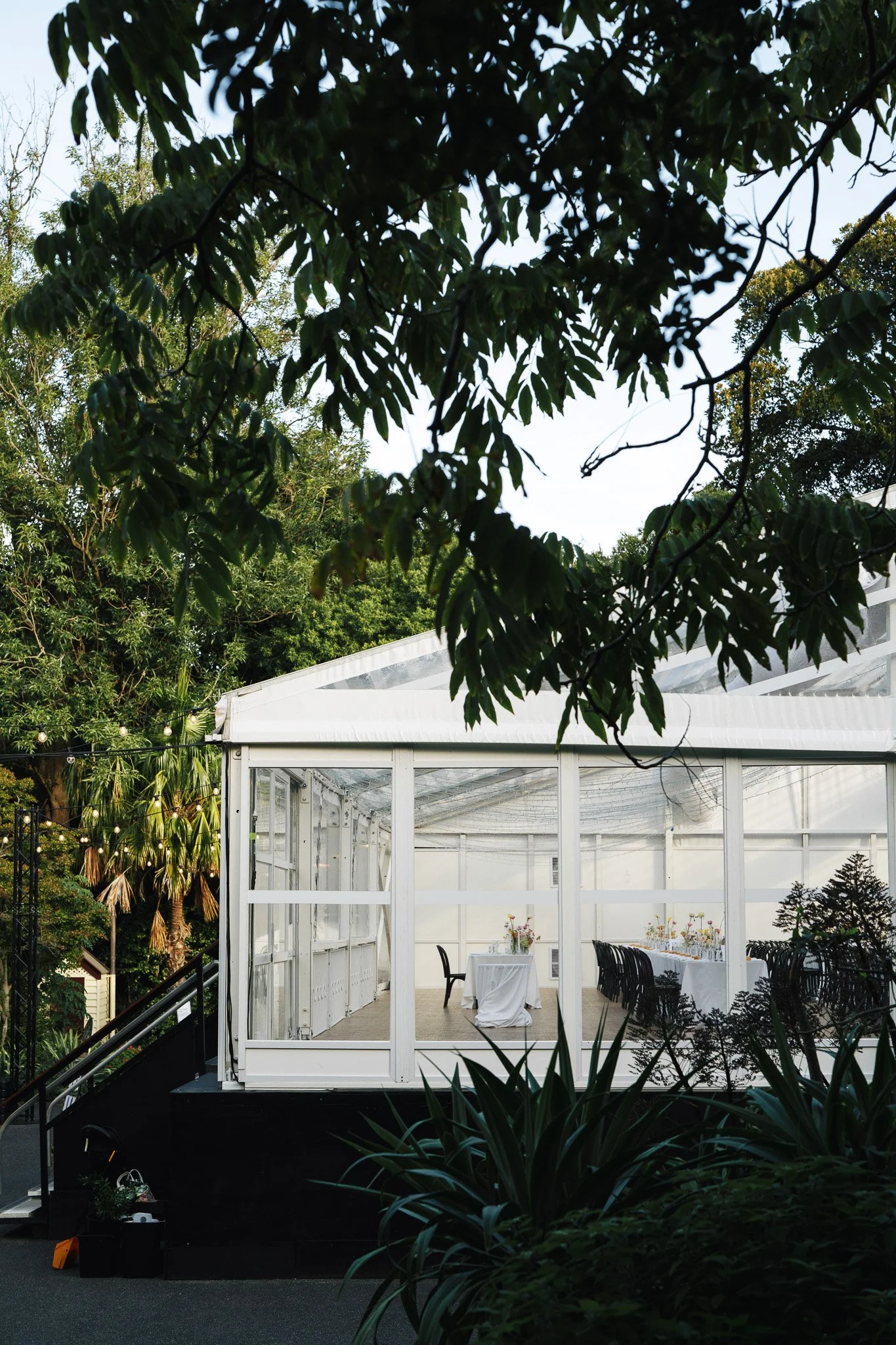 An outdoor event space with a white glass enclosure, decorated with a dining table setup, surrounded by lush green trees and foliage, with a glimpse of an elegant white tent roof and string lights.