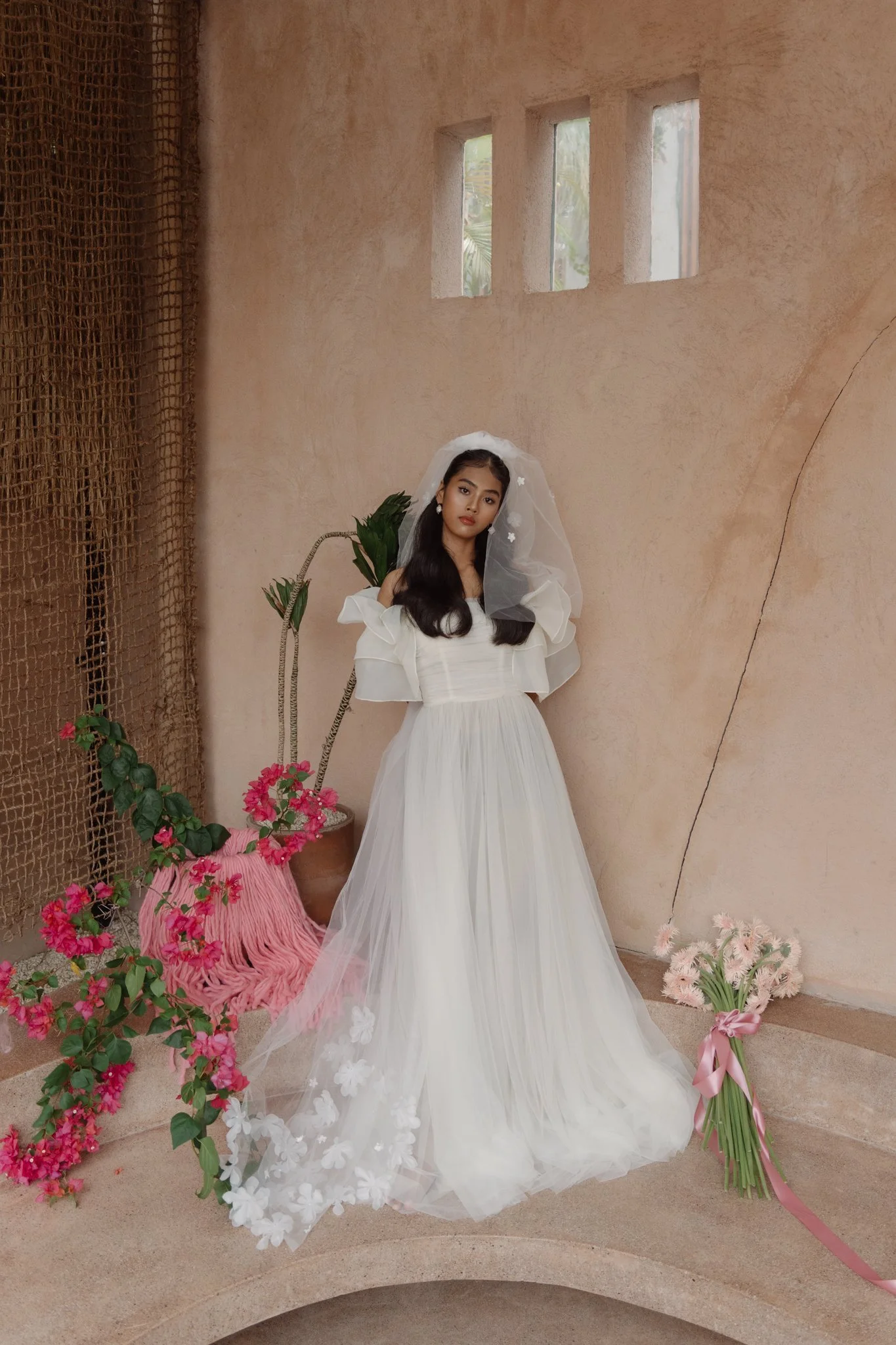 A woman in a white dress with a veil and flowers, standing against a light-colored textured wall with small windows. She is surrounded by pink and white flowers and plants.