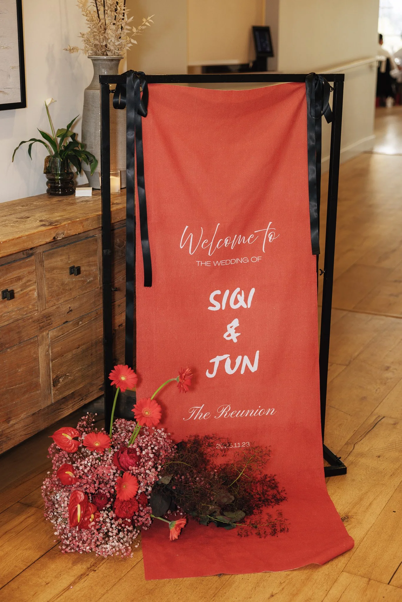 A floral arrangement with pink and red flowers and greenery in front of a red welcome sign with white text that reads "Welcome to the wedding of Sia & Jun, The Reunion" on a black metal easel. The background shows indoor decor with wooden furniture a