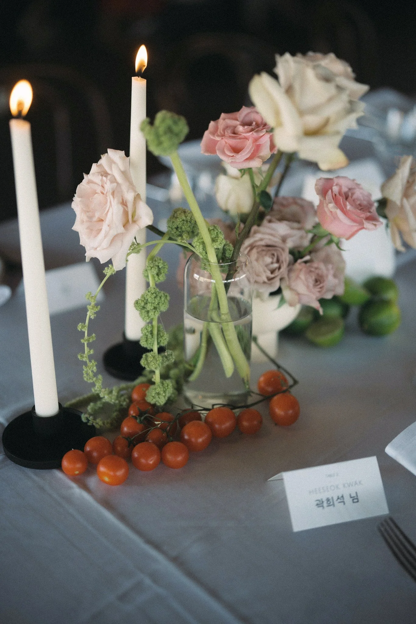 A table centerpiece featuring pink and white roses in a glass vase, surrounded by green foliage, orange cherry tomatoes, and two white candles in black candleholders, with a blurred background and a small white card with text.