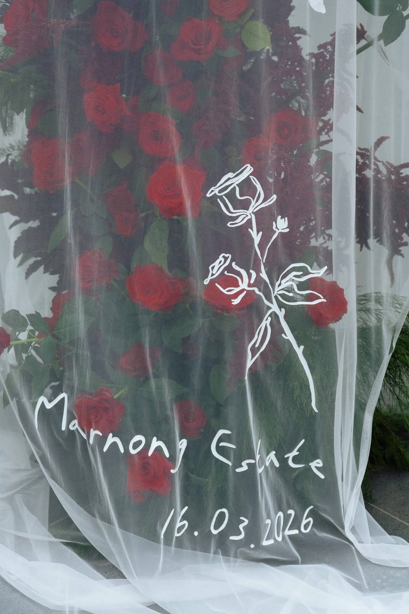 Vase of red roses with a white outline drawing of a single flower and the words 'Memoria Estate' and the date 16.03.2026 written on a sheer fabric covering the flowers.