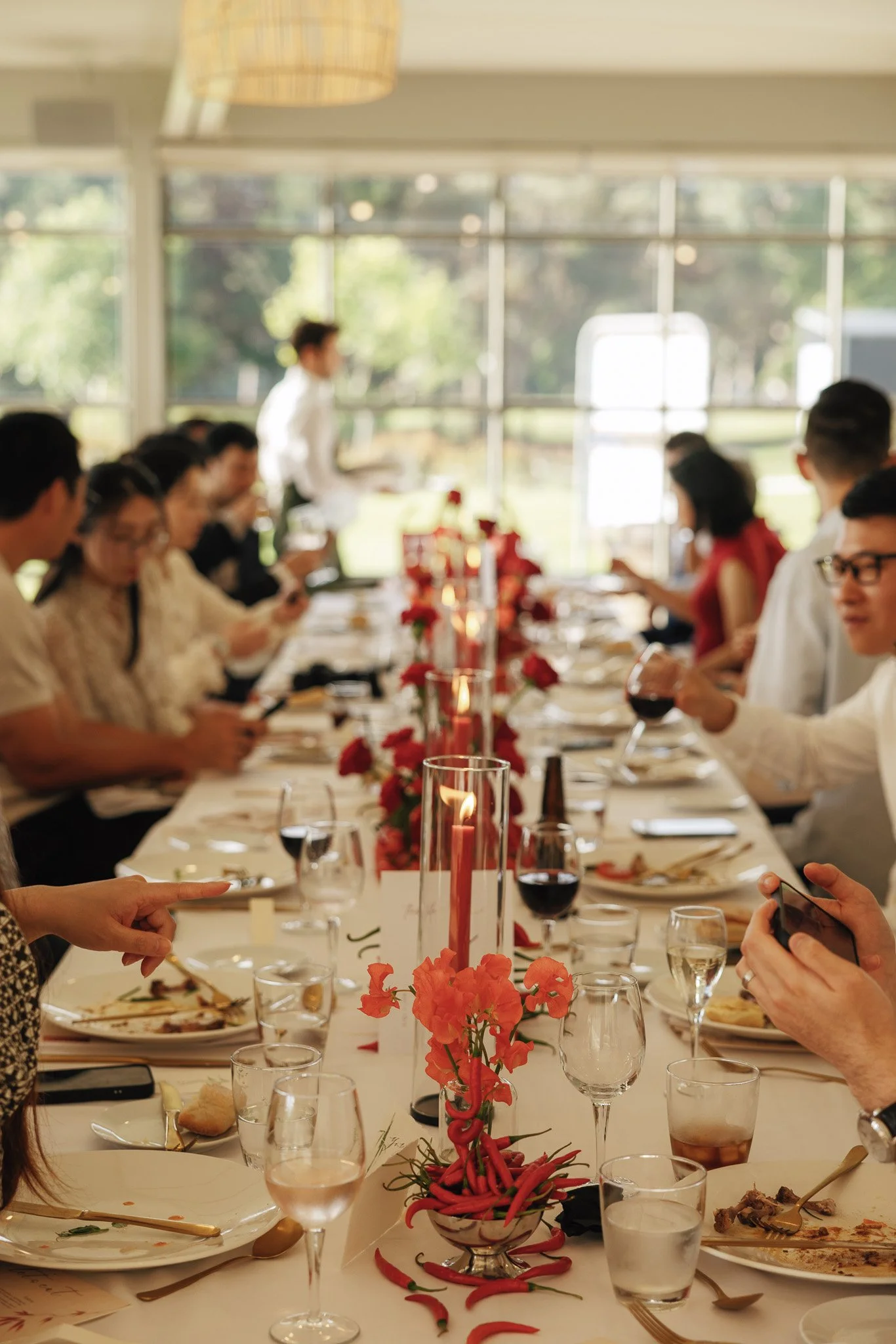 A group of people sitting at a long table during a meal, with a centerpiece of red and pink flowers and tall red candles, in a bright room with large windows showing trees outside.