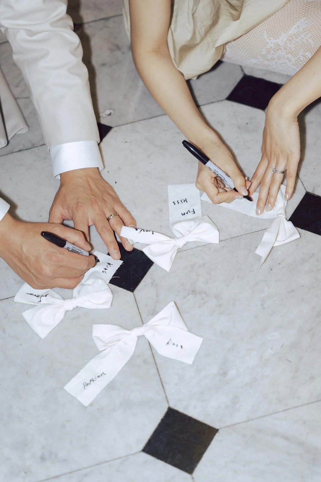 Two people are writing on white cloth bows with black markers, placing them on a tiled floor. Each bow has a handwritten name or word.
