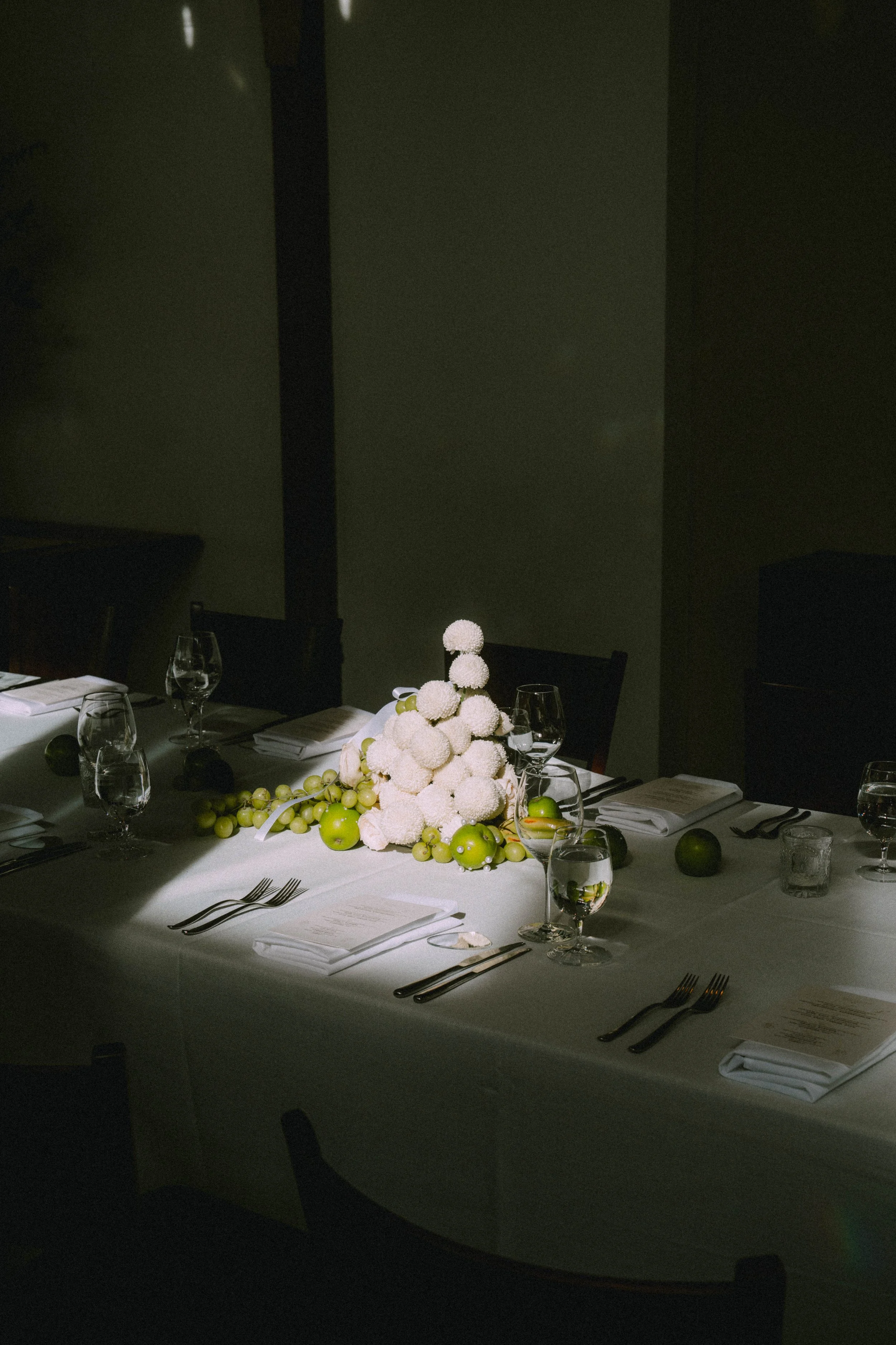 A dining table set for a meal with a white tablecloth, glasses of water, napkins, silverware, and a floral centerpiece of white spherical flowers and green apples. The room is dimly lit with a dark background.