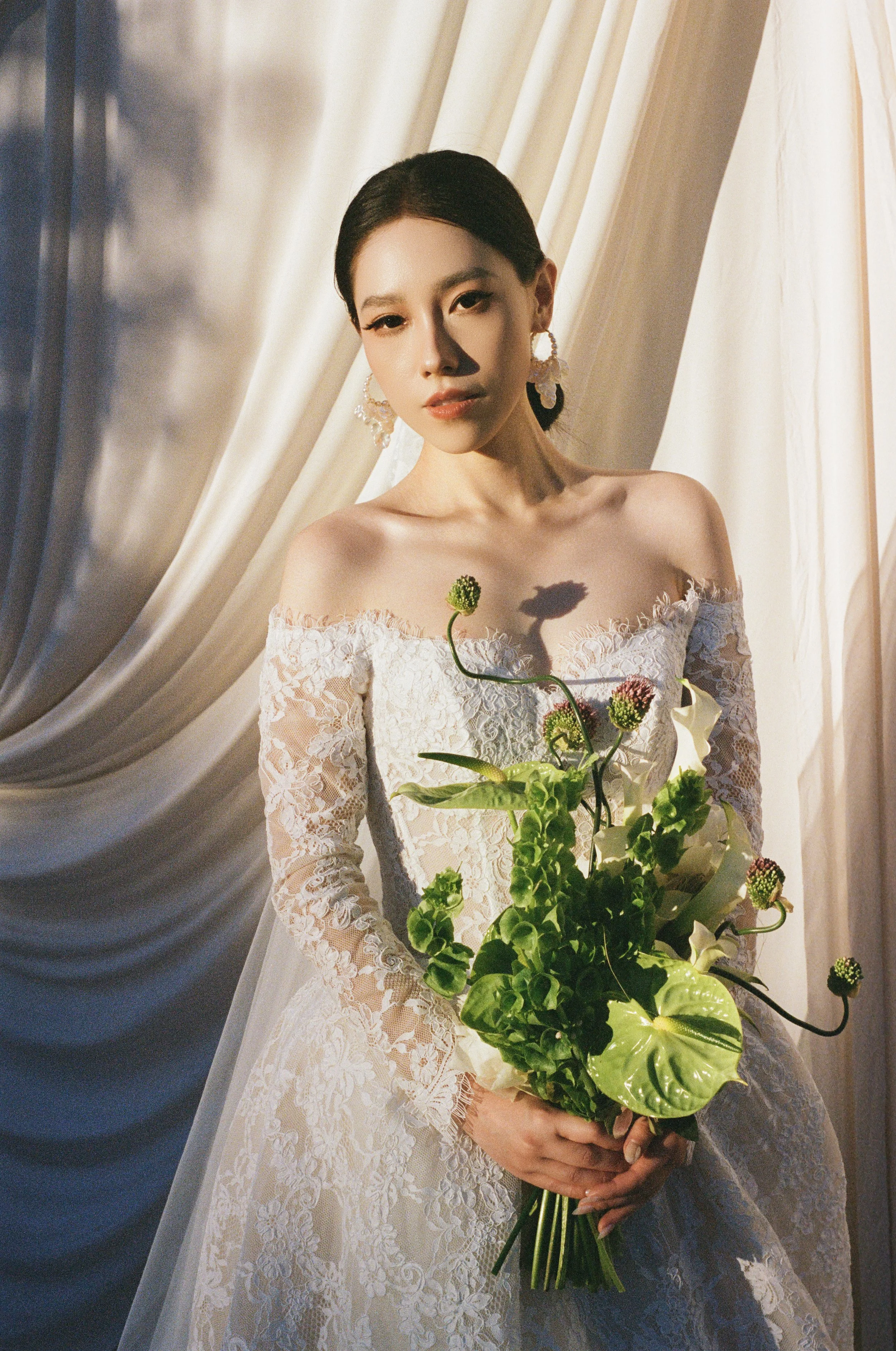 A woman in a white lace off-shoulder dress holding a bouquet of green and white flowers standing in front of cream-colored drapes.