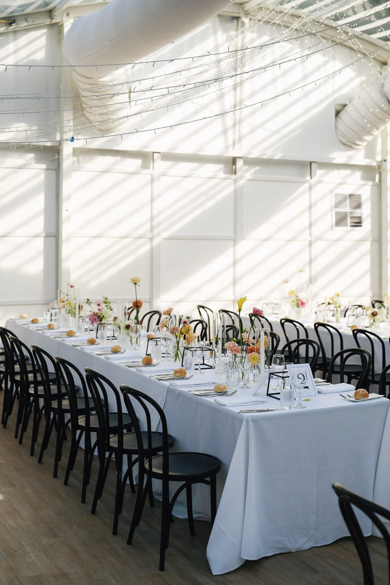 A long banquet table set for a formal event in a bright, airy space with white tablecloths, floral centerpieces, place settings, and black chairs, with sunlight streaming through a glass ceiling and string lights overhead.