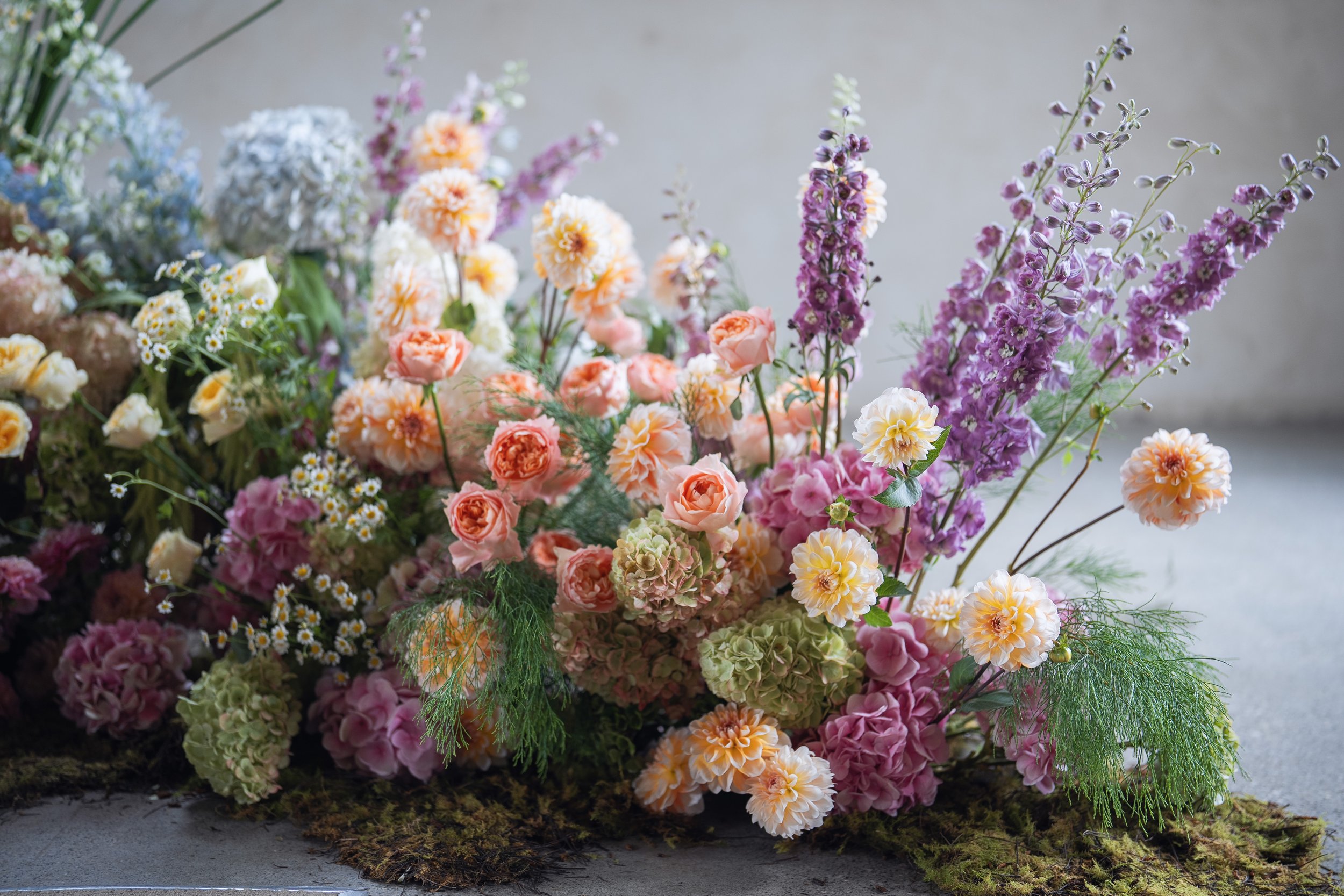 A colorful bouquet of various flowers including roses, daisies, and lavender in soft pastel shades arranged on a surface with moss and greenery.