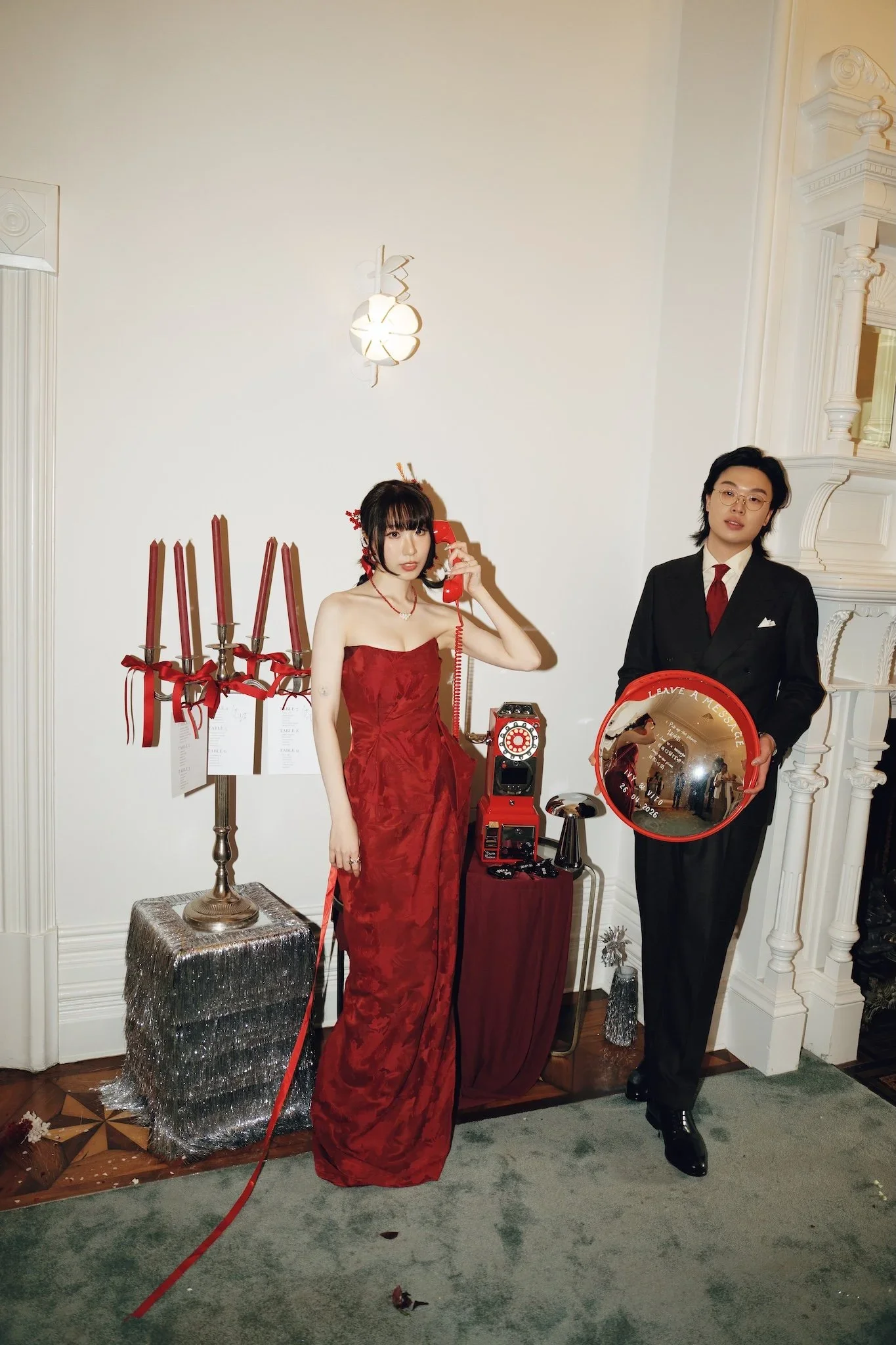 Two people standing indoors near a white wall, with one person dressed in a red gown and the other in a black suit holding a red object. There is a table with a silver-covered tablecloth and a candelabrum with red candles on the left. The scene appea
