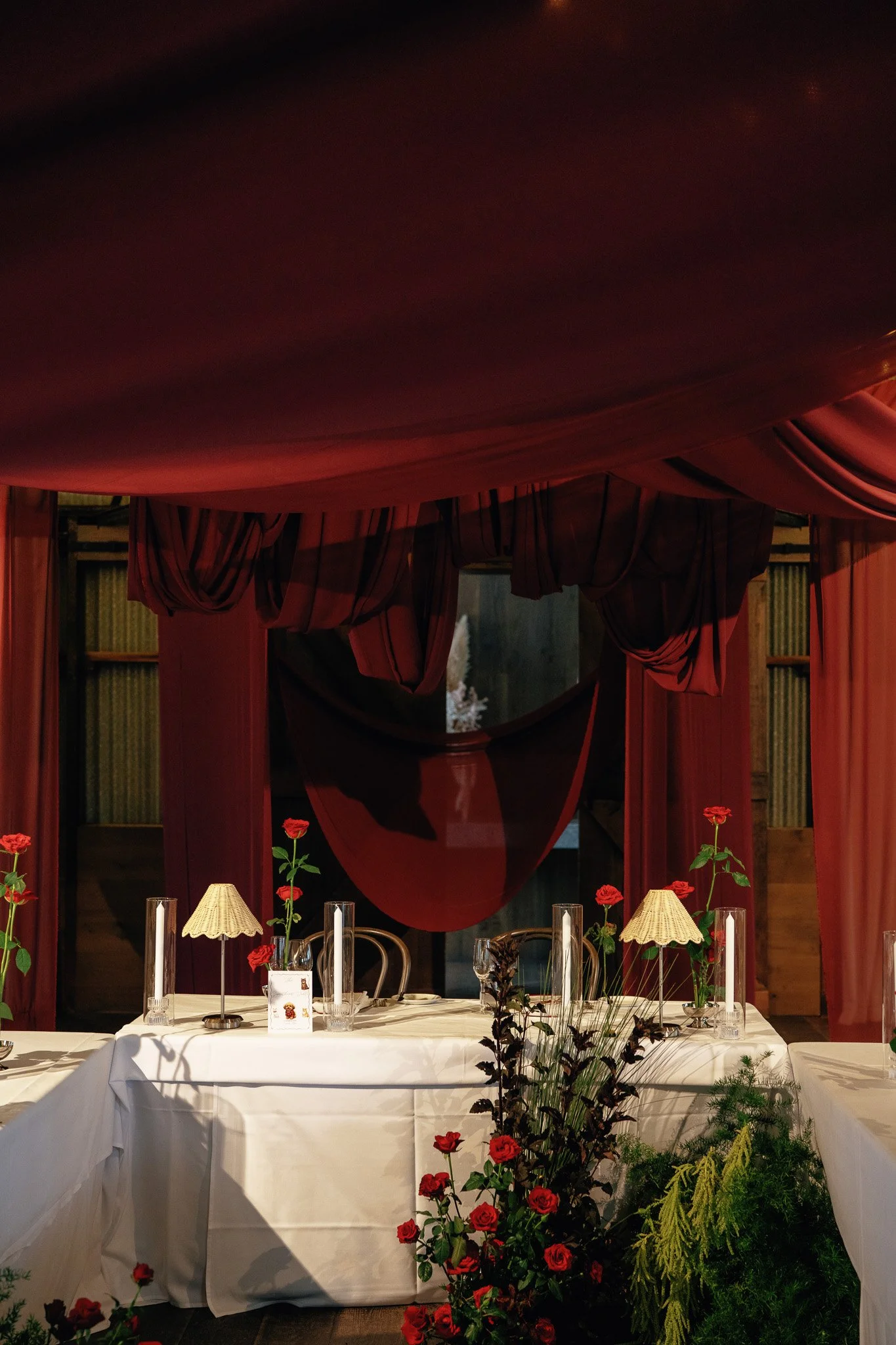 Decorated dining table with white tablecloth, red roses, candles, and small lamps. Large red drapes with a canopy or bed-like structure above, and shelves with books in the background.
