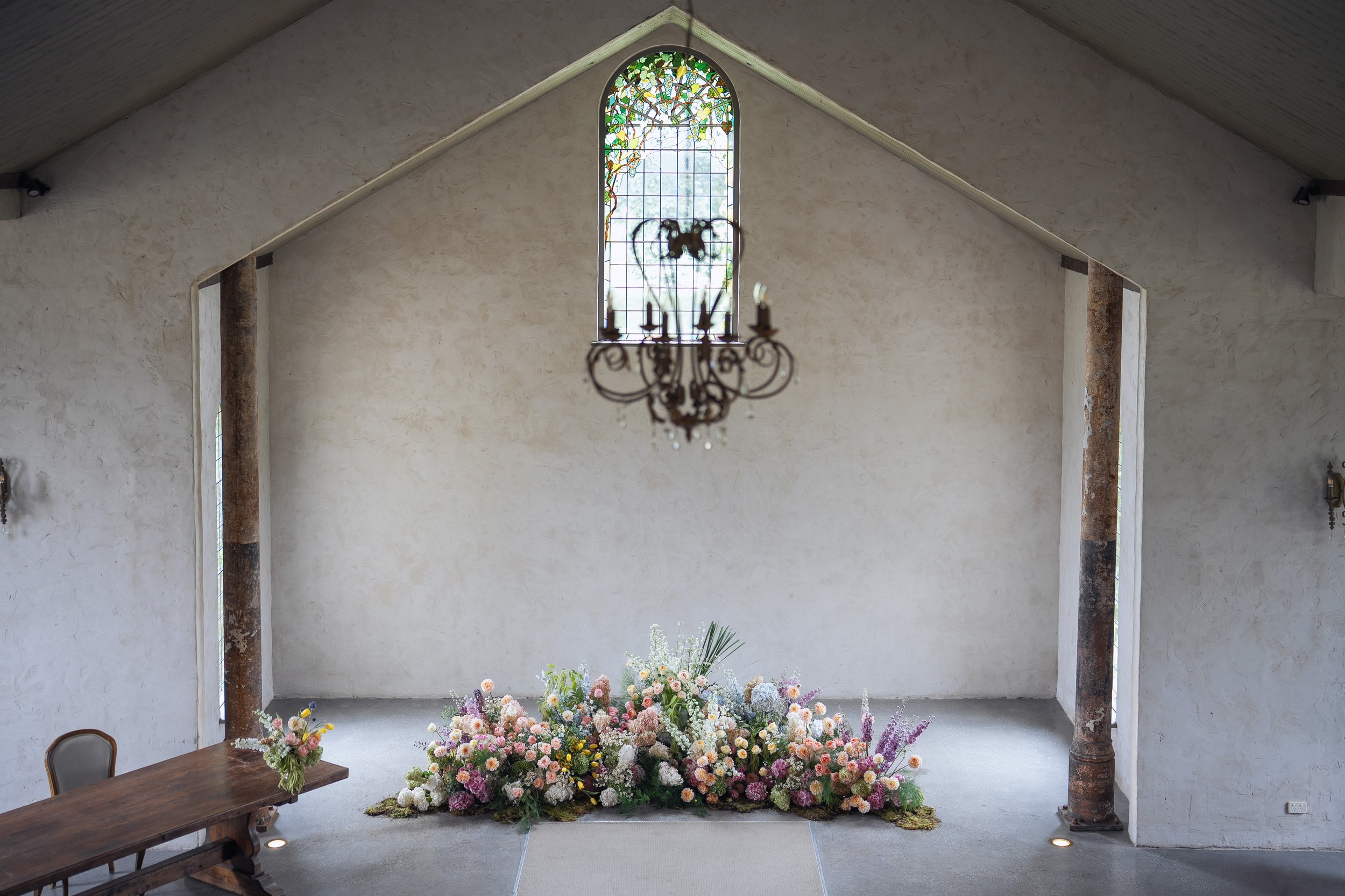 Interior of a chapel with a large stained glass window, a chandelier hanging from the ceiling, and a floral arrangement on the floor in front of the wall. There are wooden beams supporting the ceiling.