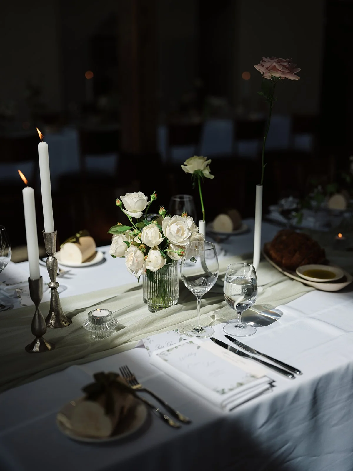 A formal dining table decorated with white roses in a glass vase, tall white candles, and glassware, set for a special occasion.