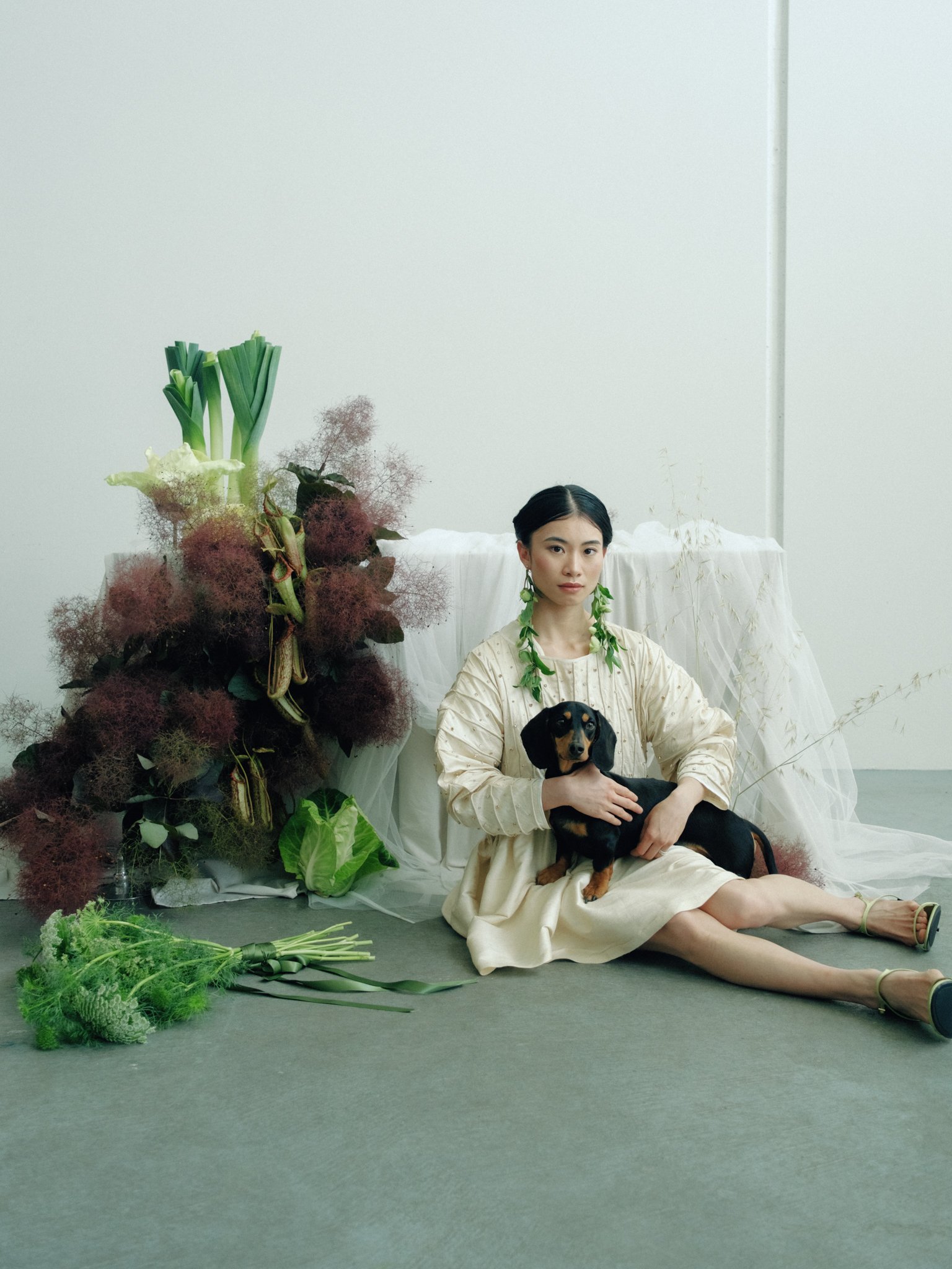 Woman sitting on the floor in front of a white backdrop, holding a black and tan dachshund dog, surrounded by various green and reddish plants and flowers.